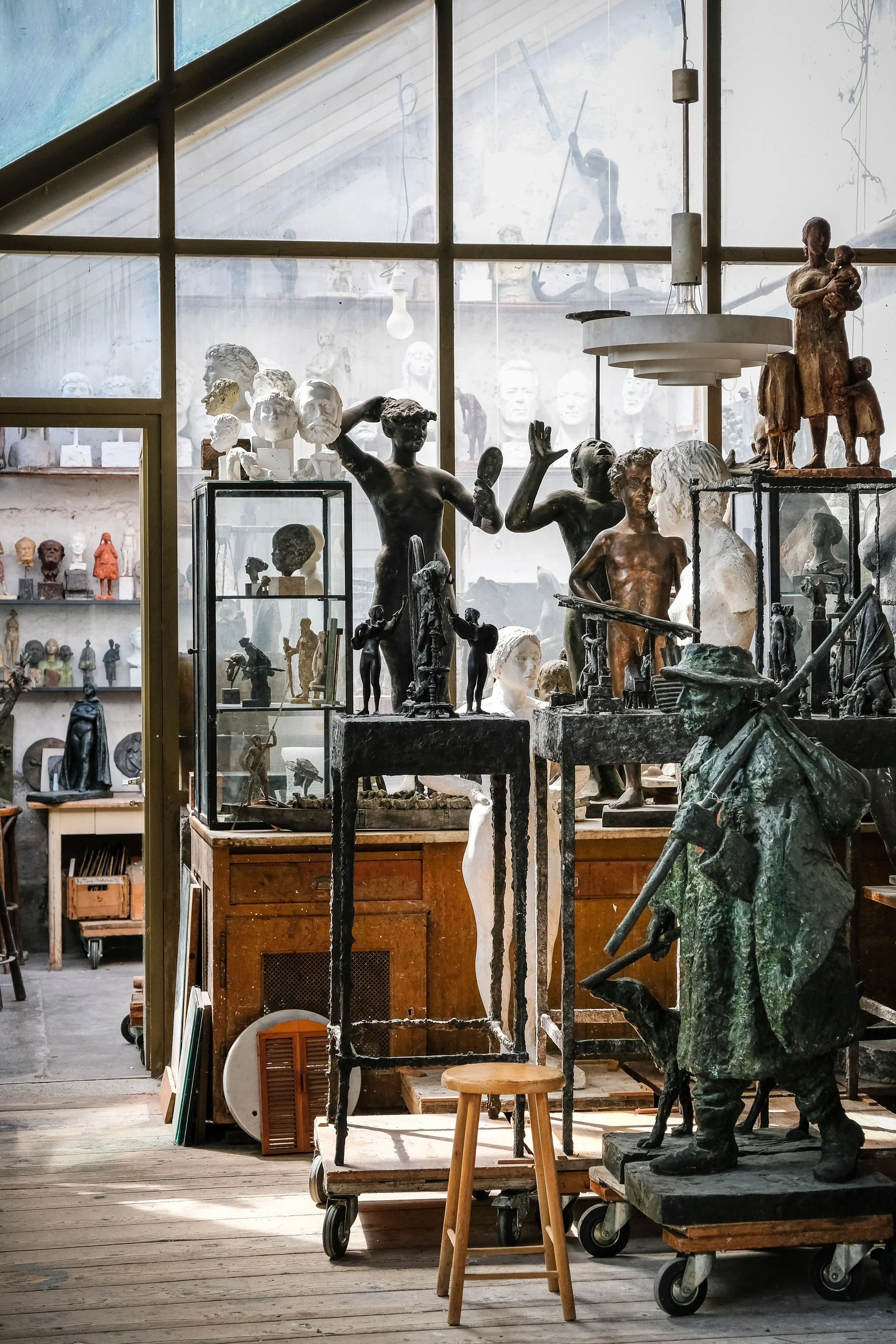 Blick in ein Kunstrestaurationsatelier mit Skulpturen und Figuren aus Bronze und anderen Materialien, auf Tischen und Rollwagen, hinter einer großen fensterartigen Wand.
