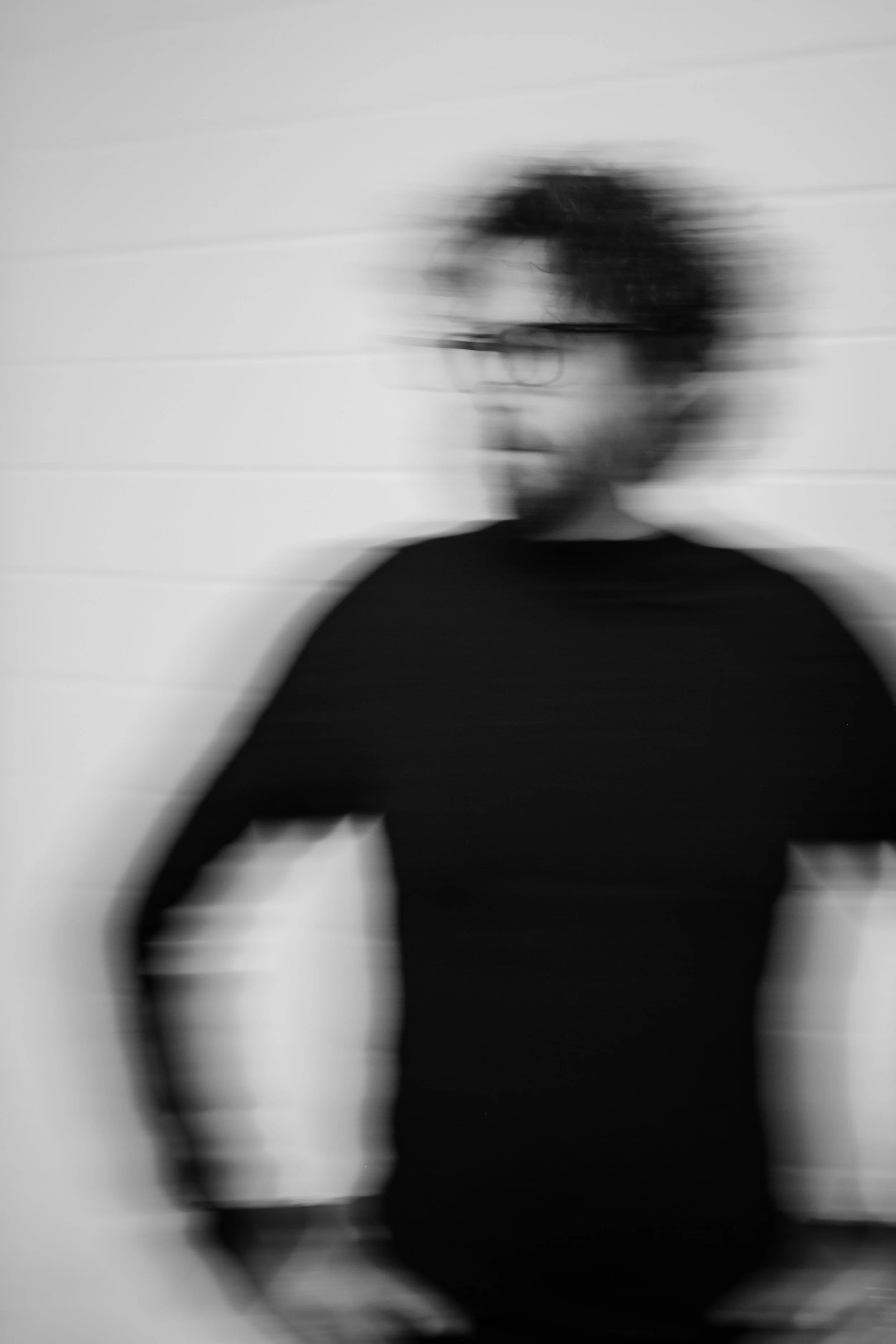 Eine verschwommene Schwarz-Weiß-Fotografie eines Mannes mit Locken, Brille und dunklem Oberteil, vor weißer Wand mit horizontalen Linien.