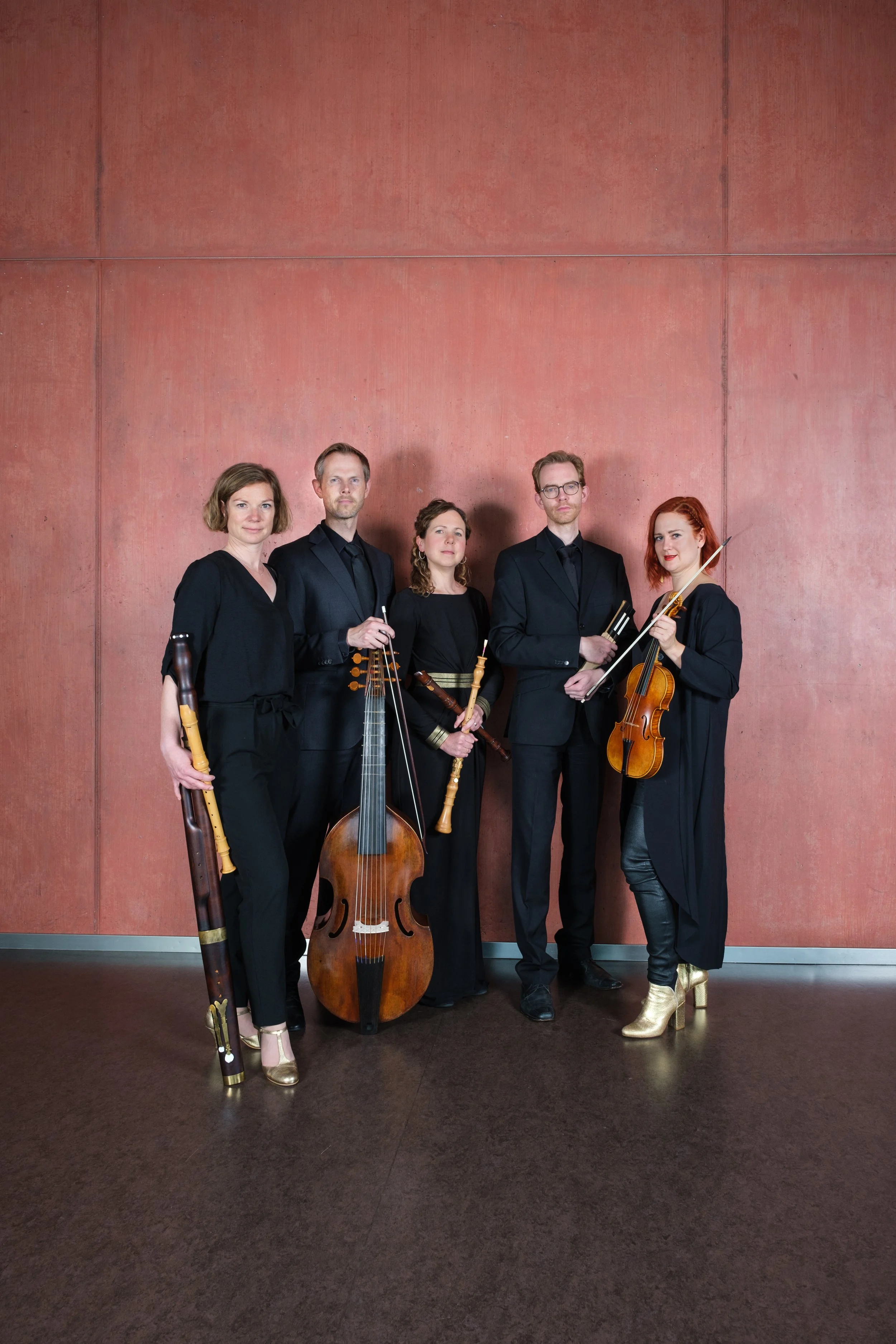 Ein klassisches Kammerorchester mit fünf Mitgliedern, das verschiedene Instrumente hält, vor einer roten Wand.
