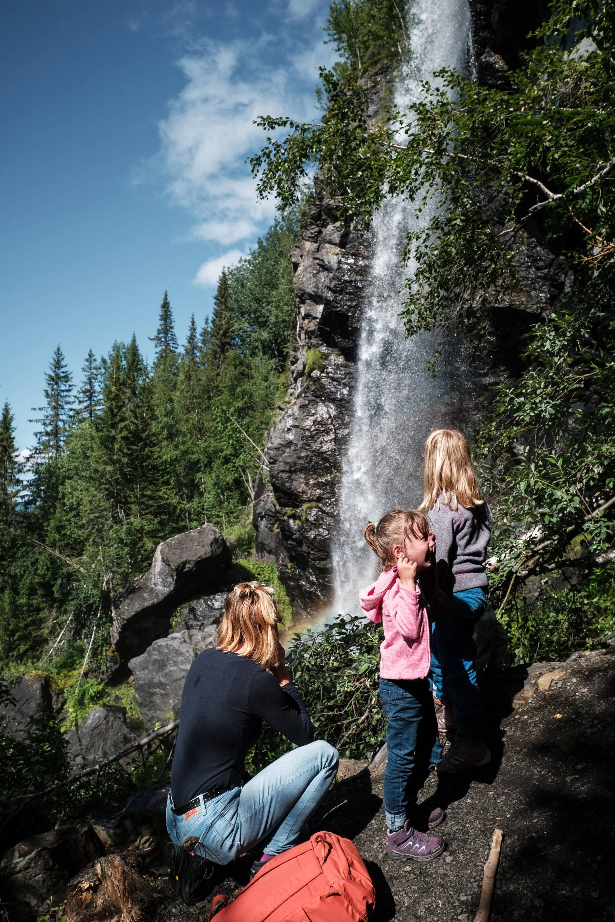 Ein Mädchen sitzt auf einem Felsen und beobachtet eine Gruppe von Kindern, die vor einem Wasserfall stehen, umgeben von grünen Bäumen unter einem blauen Himmel.