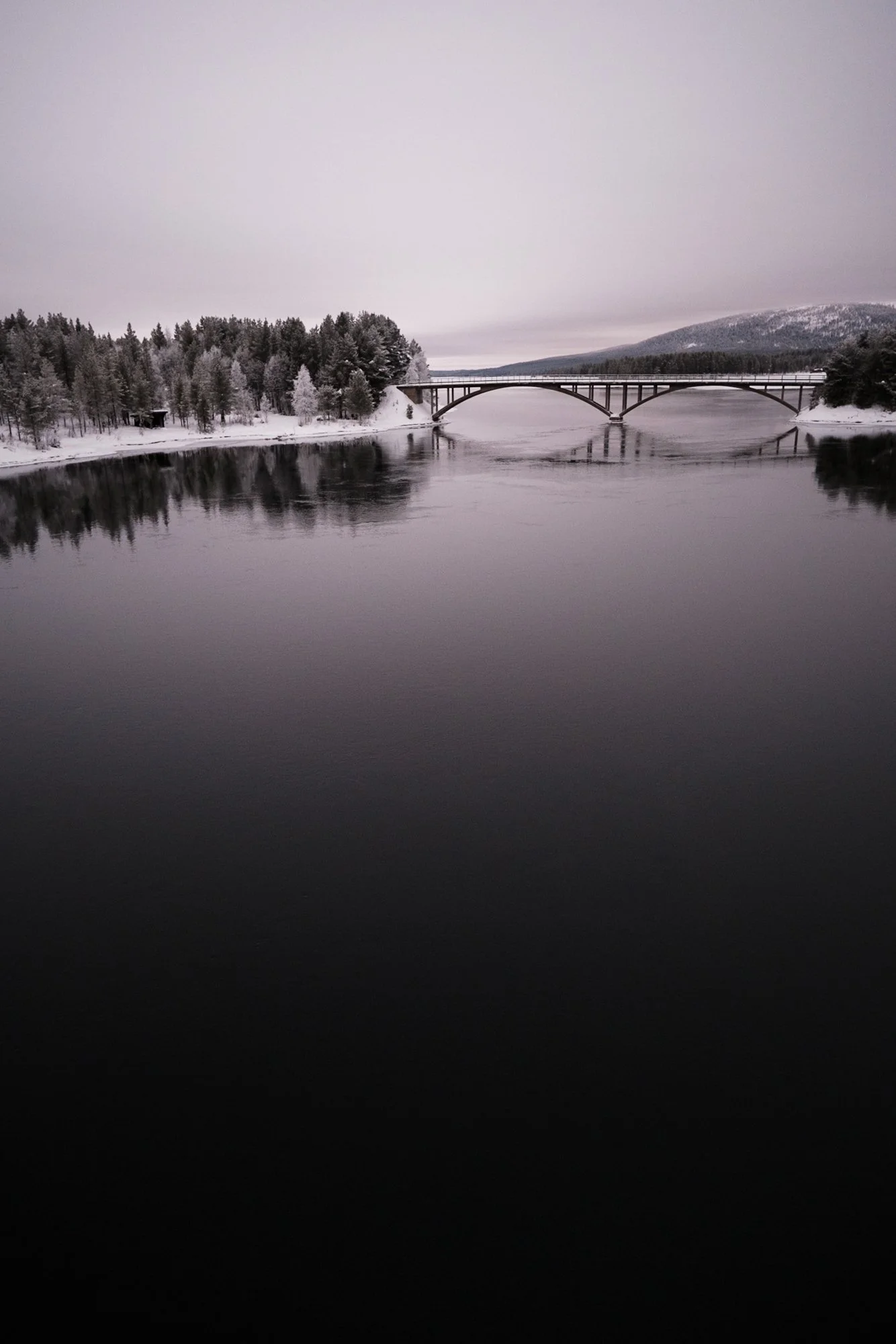 Ein Fluss im Winter mit spiegelndem Wasser, umgeben von schneebedeckten Bäumen und einem Brücke im Hintergrund, grauer Himmel.