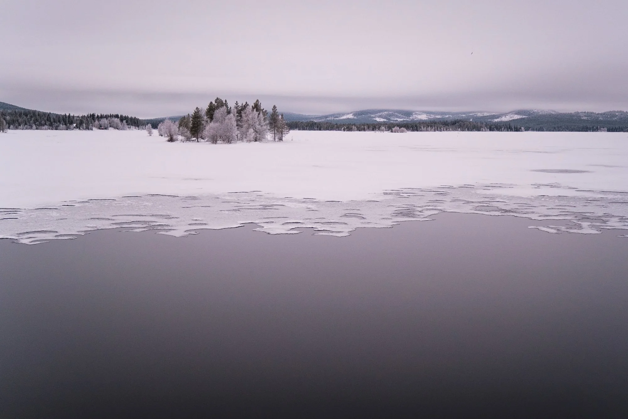 Ein verschneiter, gefrorener See mit Eisflecken und Bäumen im Hintergrund, unter einem bedeckten Himmel.
