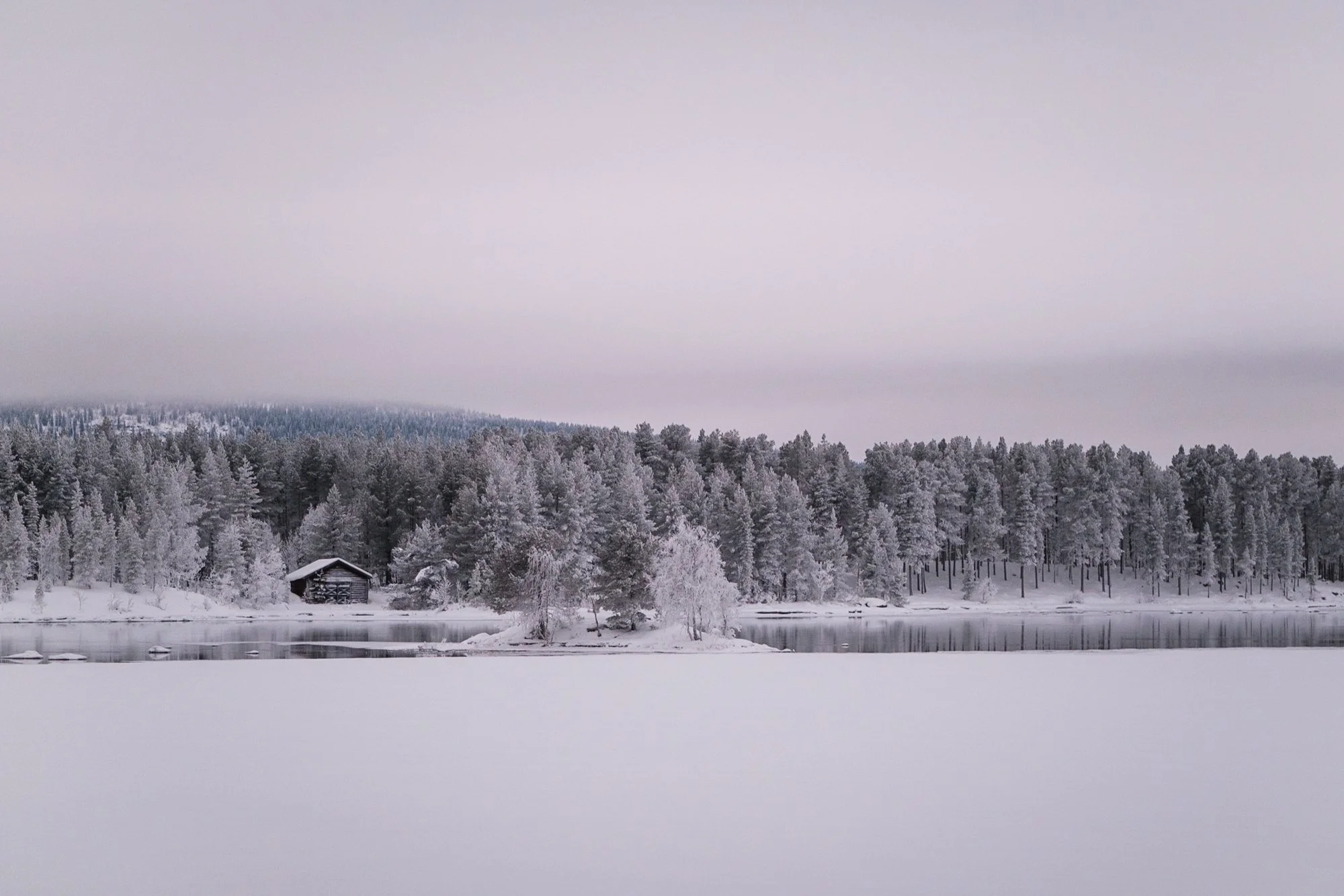 Schneebedeckter See mit Wald im Hintergrund und einem kleinen Holzhaus am Ufer, alles von Schnee bedeckt im Winter.