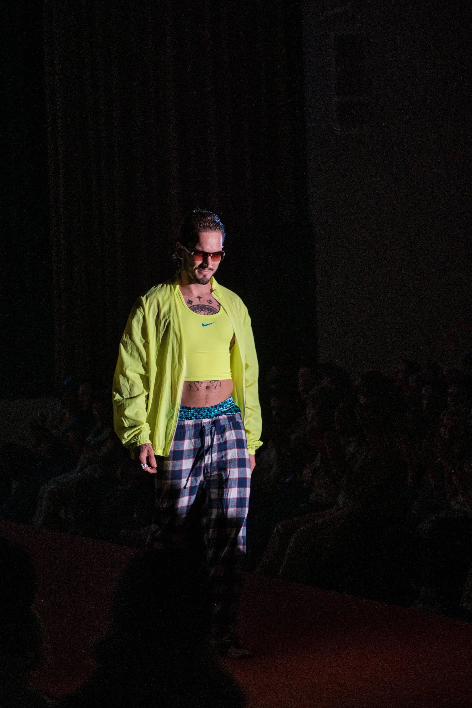 Mann auf Laufsteg in dunklem Raum, trägt neon gelbe Jacke, Crop Top, gemusterte Hosen, Sonnenbrille.