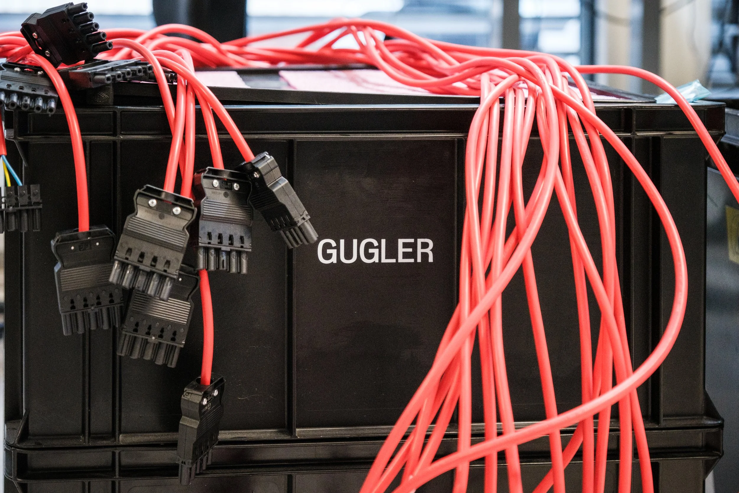 Schwarzer Kasten mit dem Label 'GUGLER' und roten Elektrokabeln, die darüber und hinaus verlaufen.
