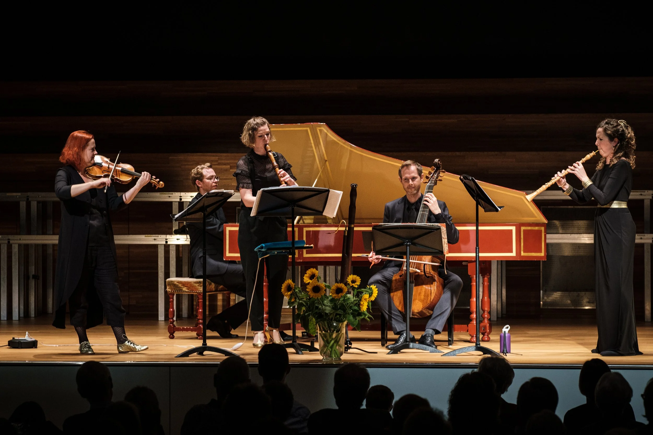 Fünf Musiker spielen auf einer Bühne mit einem goldgelben Klavier im Hintergrund, während sie Instrumente wie Violine, Blockflöte, Cello und Mandoline spielen, vor Publikum.