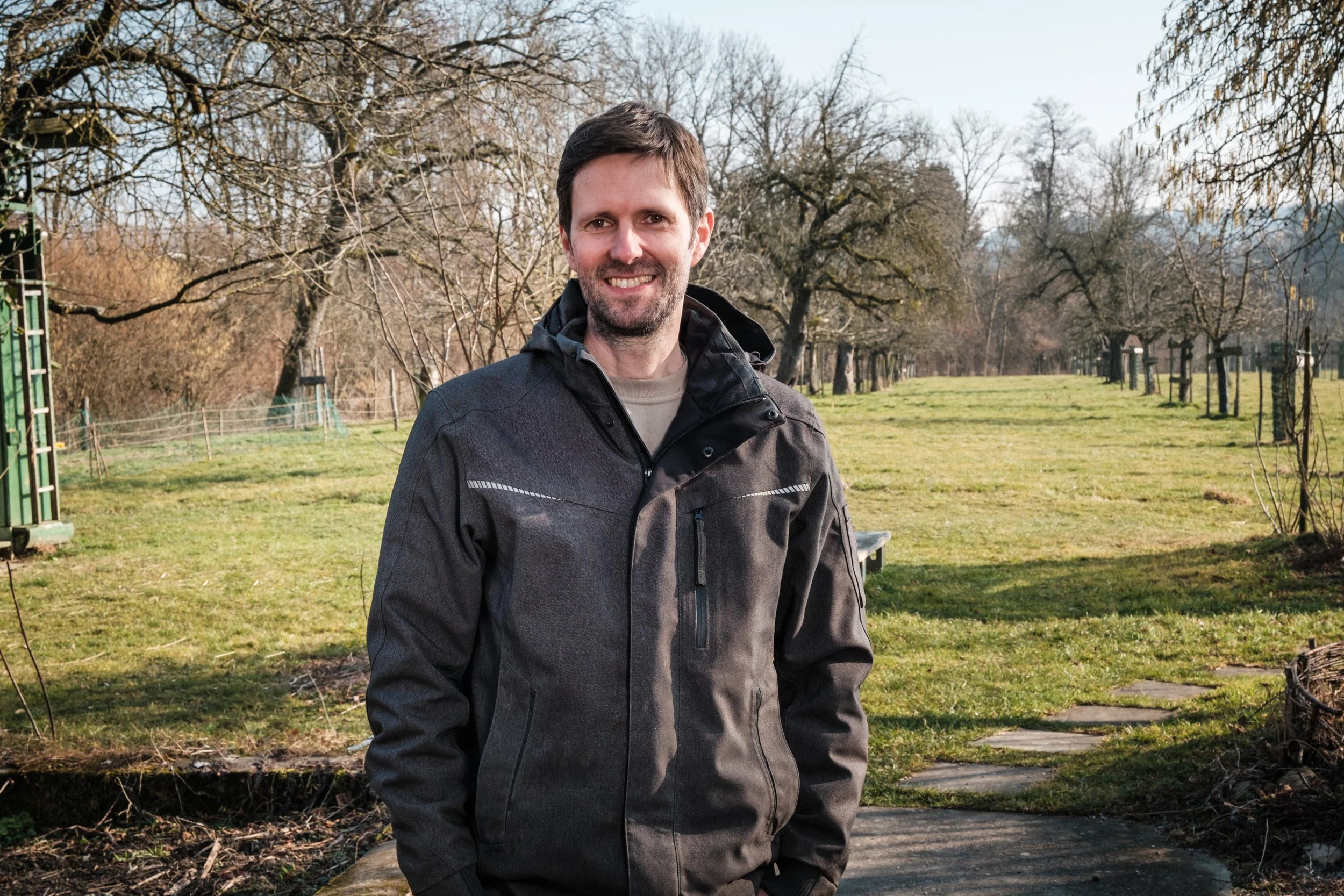 Ein lachender Mann steht im Freien auf einem Weg, im Hintergrund sind Bäume ohne Blätter und eine grüne Wiese mit Steinen und Bänken.