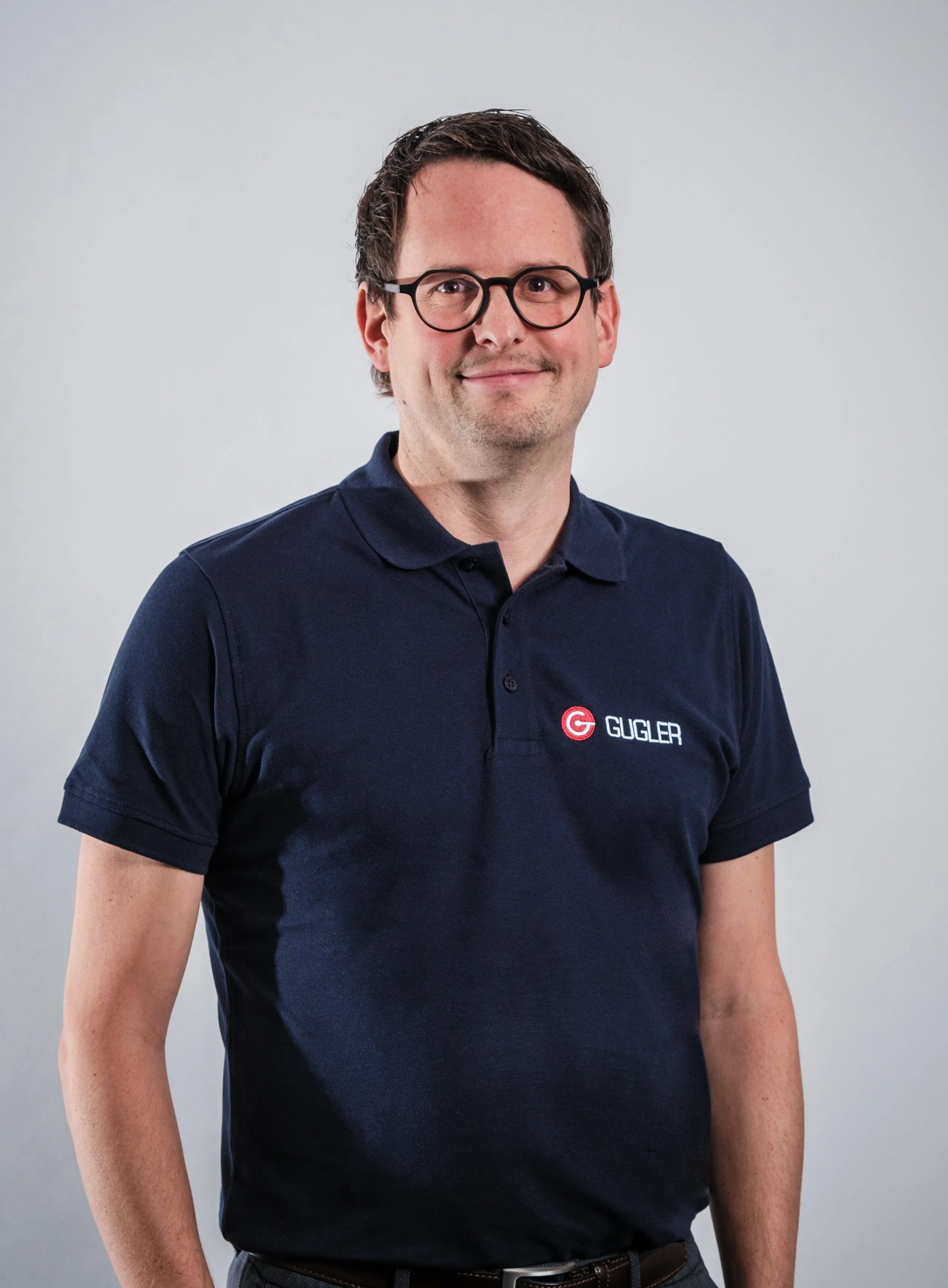Ein Mann trägt eine dunkelblaue Poloshirt mit einem Logo und dem Schriftzug 'GUGLER' auf der Brust, trägt schwarze Brille, hat dunkle, kurze Haare und steht vor einem hellgrauen Hintergrund.