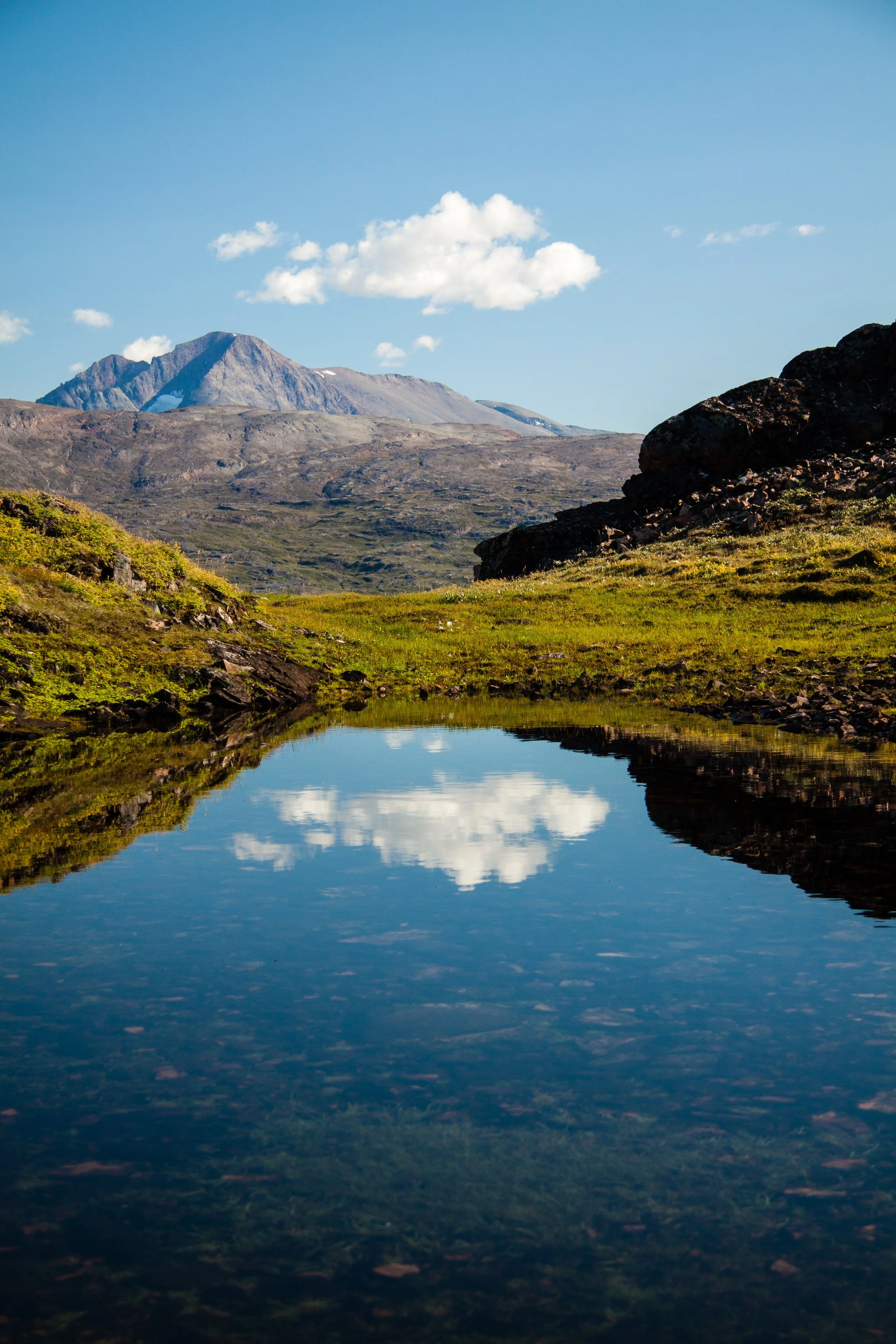 Ein Bergpanorama mit einer ruhigen Wasserfläche im Vordergrund, die den Himmel und die umliegende Landschaft spiegelt, unter einem blauen Himmel mit einigen Wolken.