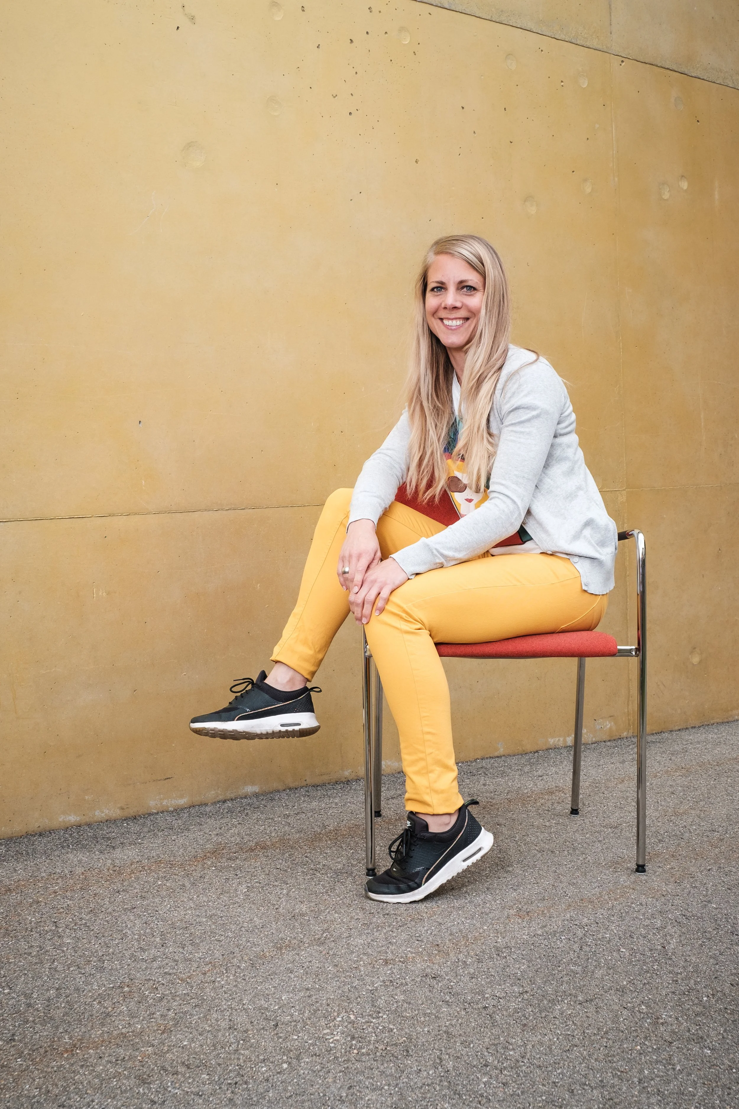 Eine lachende Frau mit langen blonden Haaren sitzt auf einem roten Stuhl vor einer gelben Wand, trägt eine graue Jacke, ein buntes T-Shirt, gelbe Hose und schwarze Turnschuhe.