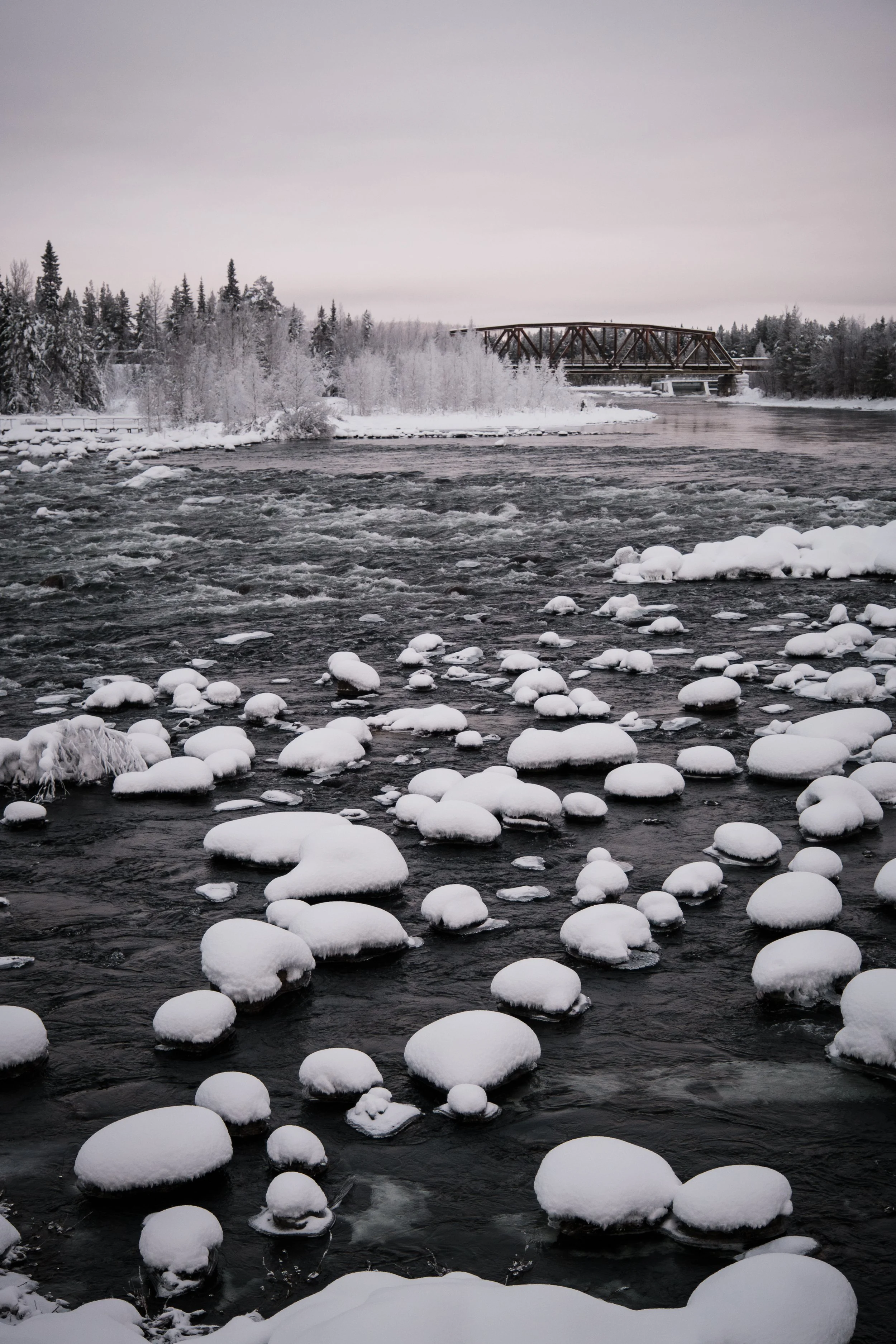 Ein verschneiter Fluss mit Eis und Schnee auf den Steinen, im Hintergrund Bäume und eine Brücke unter einem grauen Himmel.