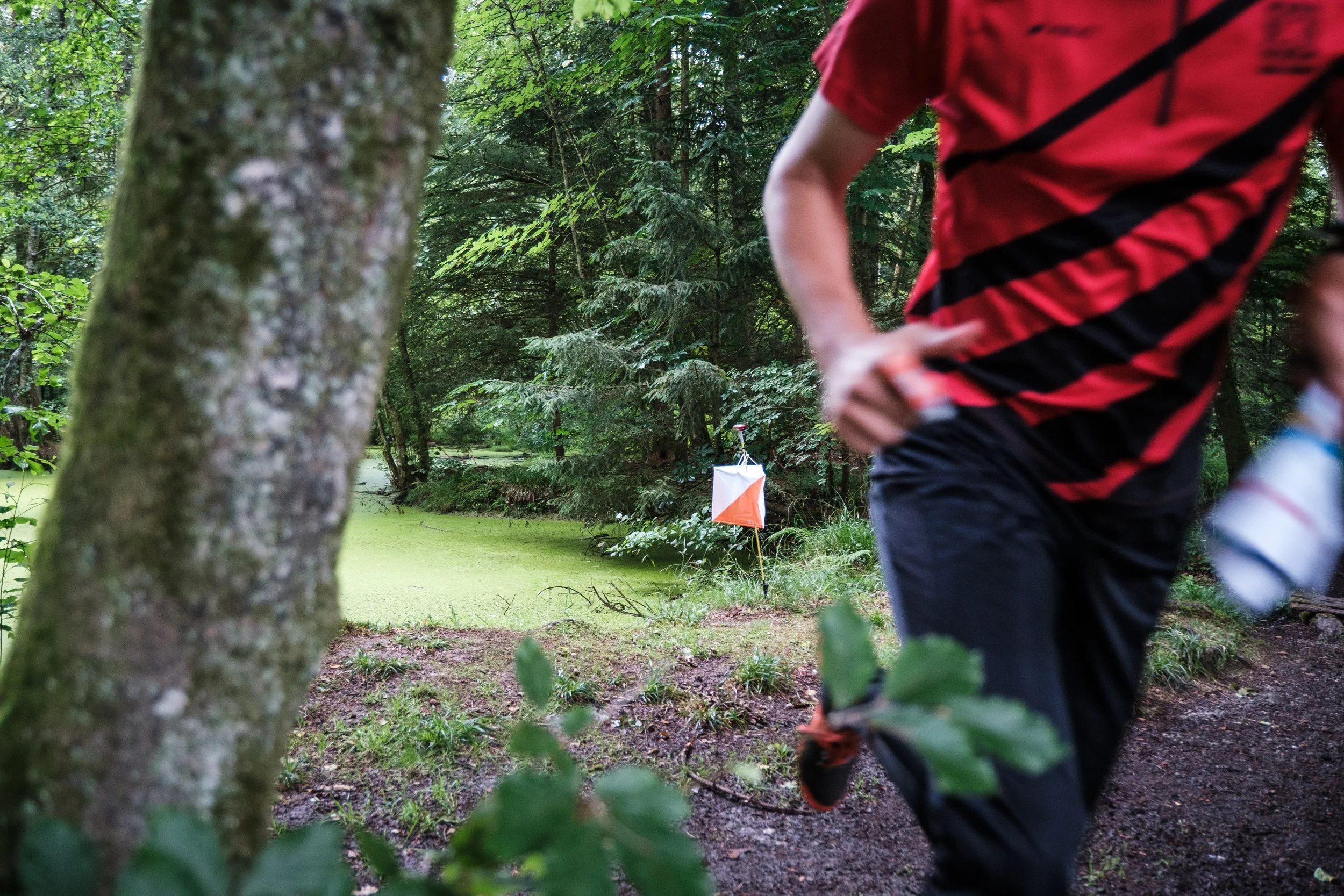 Ein Mensch in roter und schwarzer Sportkleidung, der im Wald läuft, mit einem Wasserflasche in der Hand. Im Hintergrund ist eine Boje im Wasser zu sehen, auf einem kleinen See oder Teich, mit umgebendem grünen Wald.