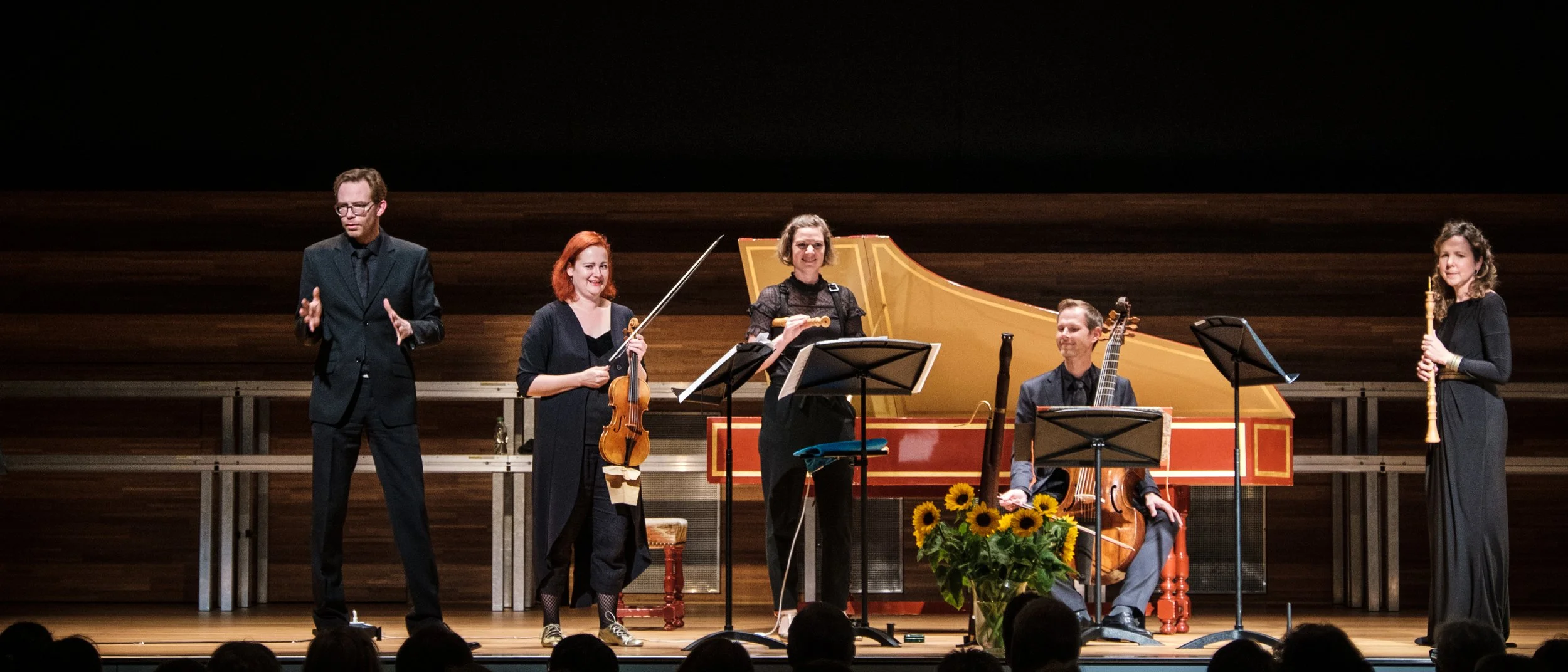 Fünf Musiker auf einer Bühne, zwei Frauen und drei Männer, mit Instrumenten wie Violine, Klavier, Cello, Flöte, Gitarre und Kontrabass, während sie ein Konzert aufführen.