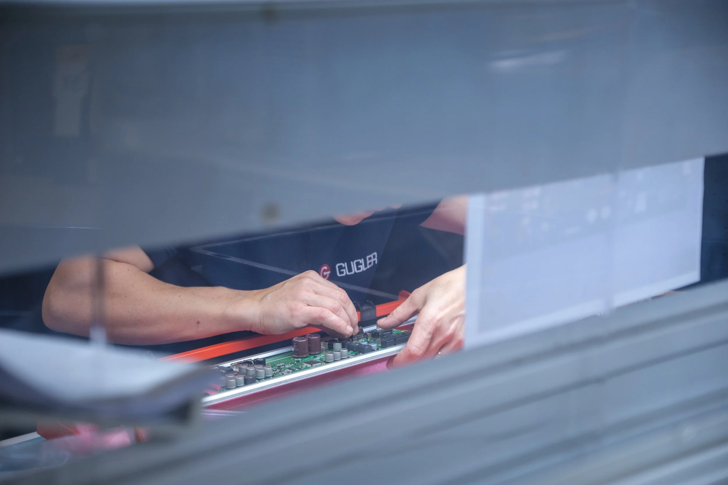 Nahaufnahme von Händen, die an einem elektronischen Bauteil auf einer Leiterplatte arbeiten, durch eine größere Metallabdeckung oder ein Gehäuse betrachtet.