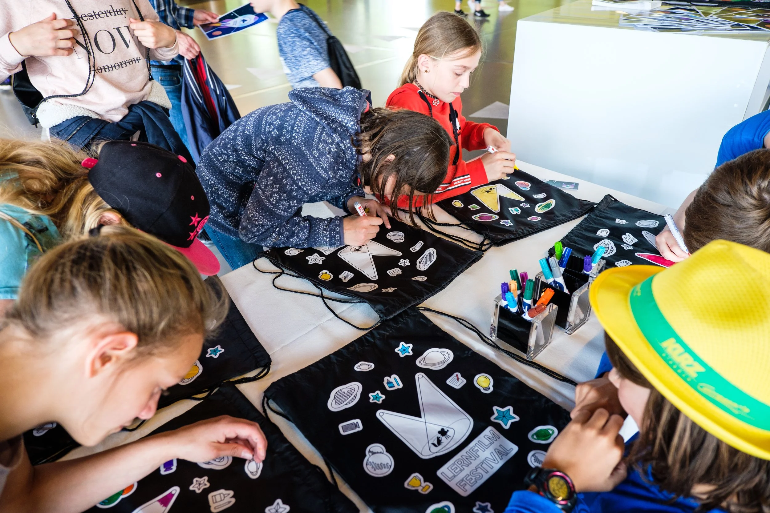 Kinder beim Basteln an einem Basteltisch mit Aufklebern und Stiften, trug symbolische schwarze Taschen mit dem Logo des Lernfilm Festivals.