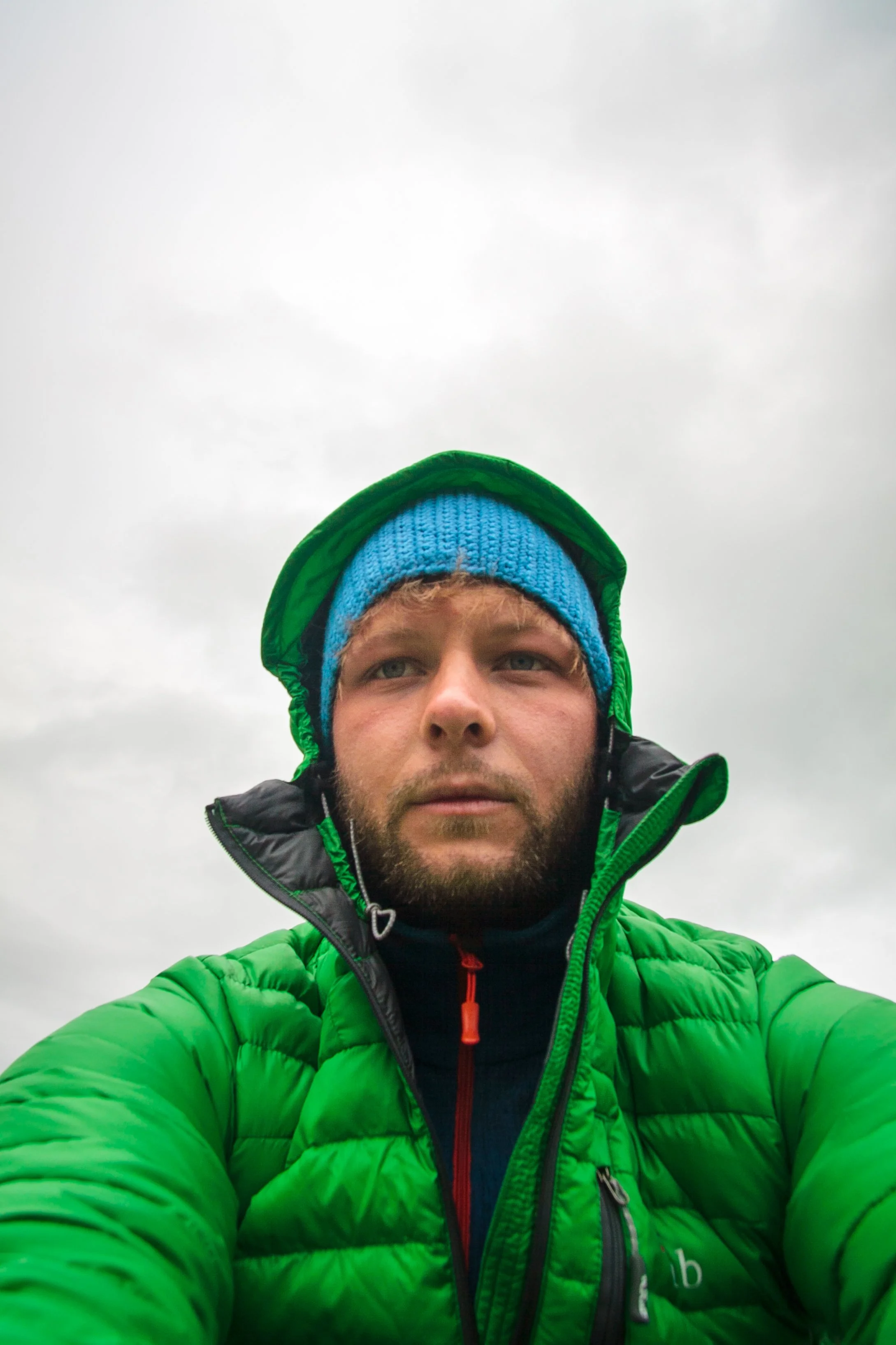 Ein Mann in grüner Jacke und blauer Mütze macht ein Selfie im Freien bei bewölktem Himmel.