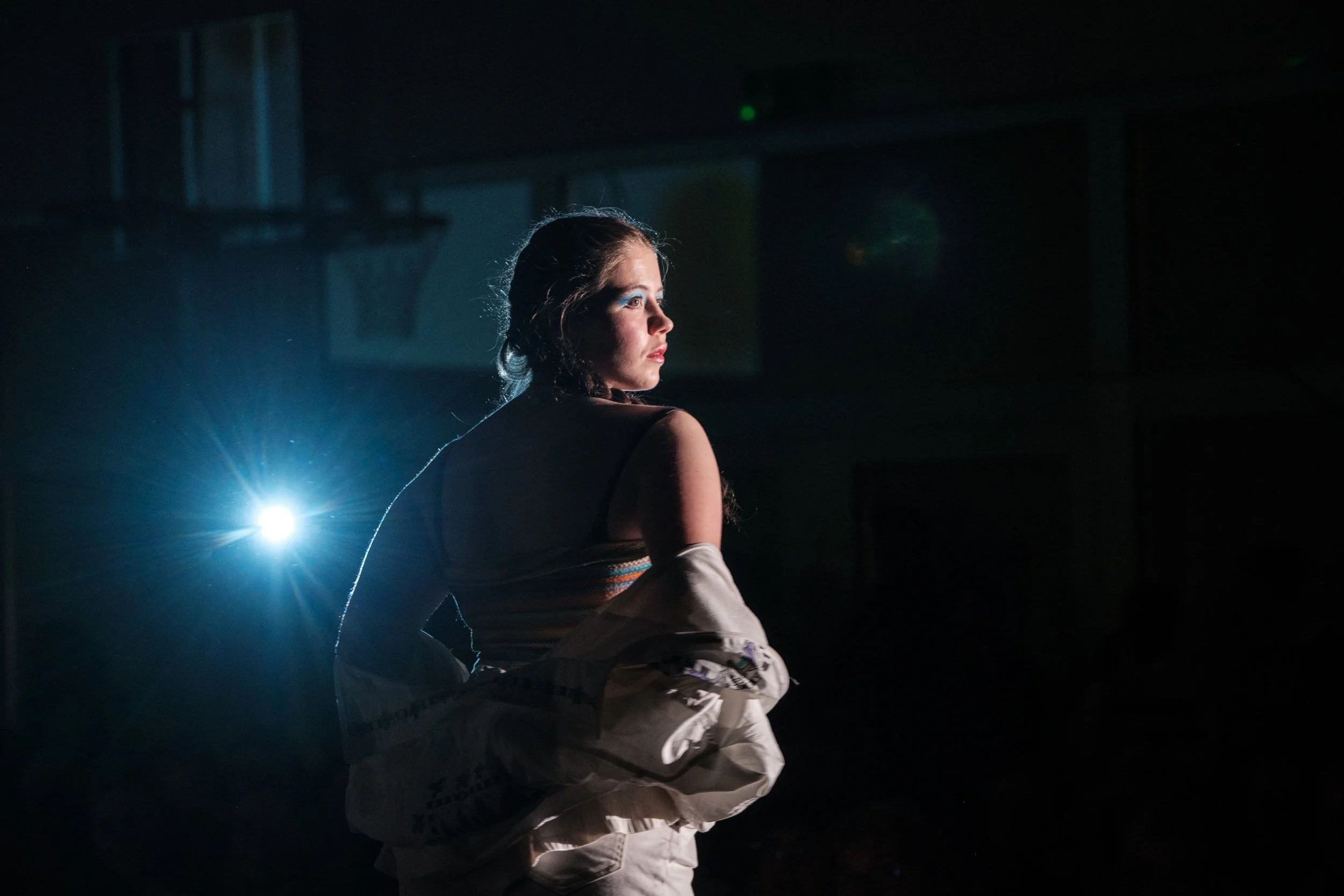 Junge Frau in dunklem Raum mit Lichtquelle im Hintergrund, seitlicher Blick, trägt einen farbigen Oberteil und eine Jacke in der Hand.