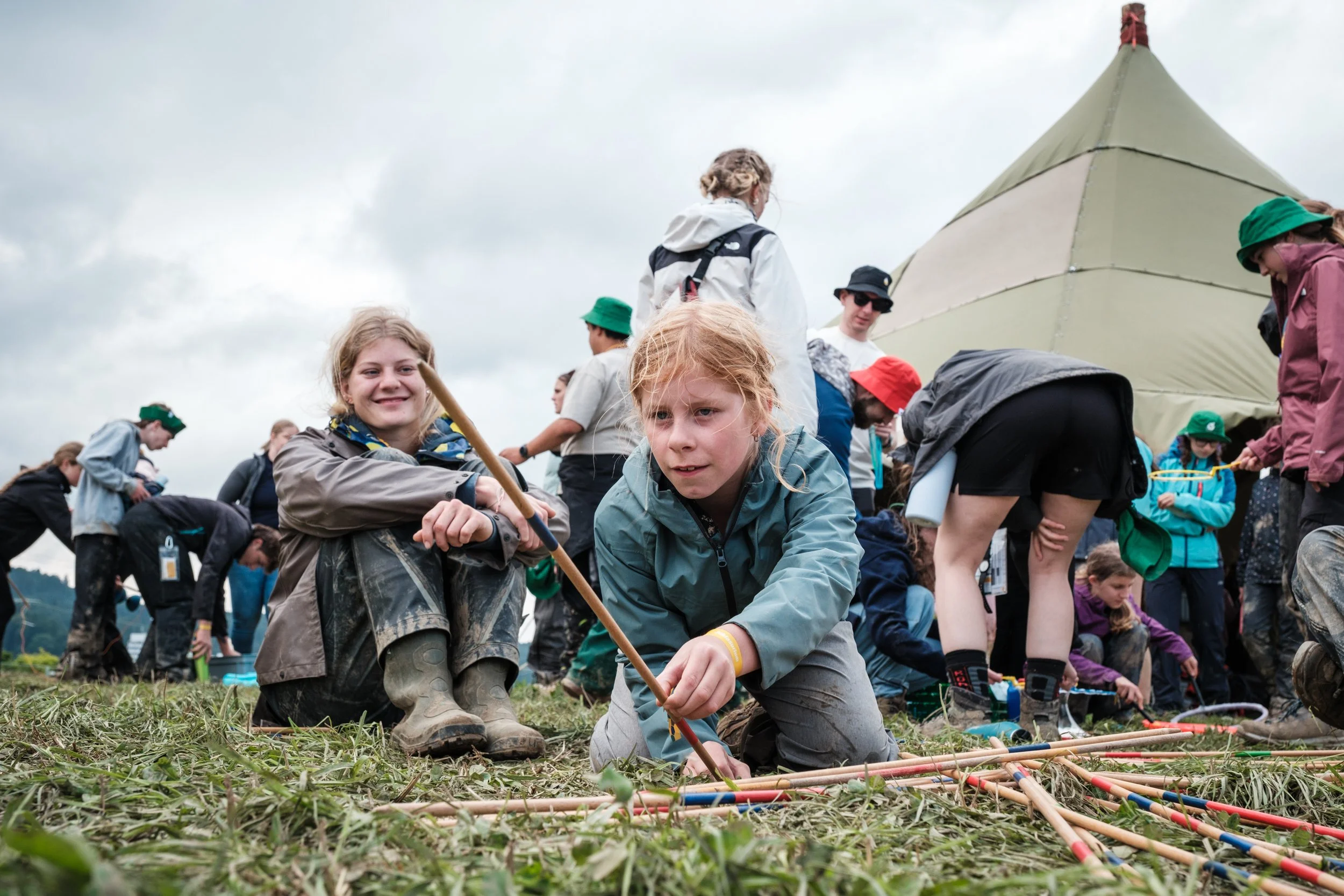 Viele Kinder beim Picknick im Freien, einige arbeiten an einem Projekt mit Rohren oder Stöcken, während sie auf dem Gras sitzen, im Hintergrund ein Zelt und eine Gruppe von Menschen