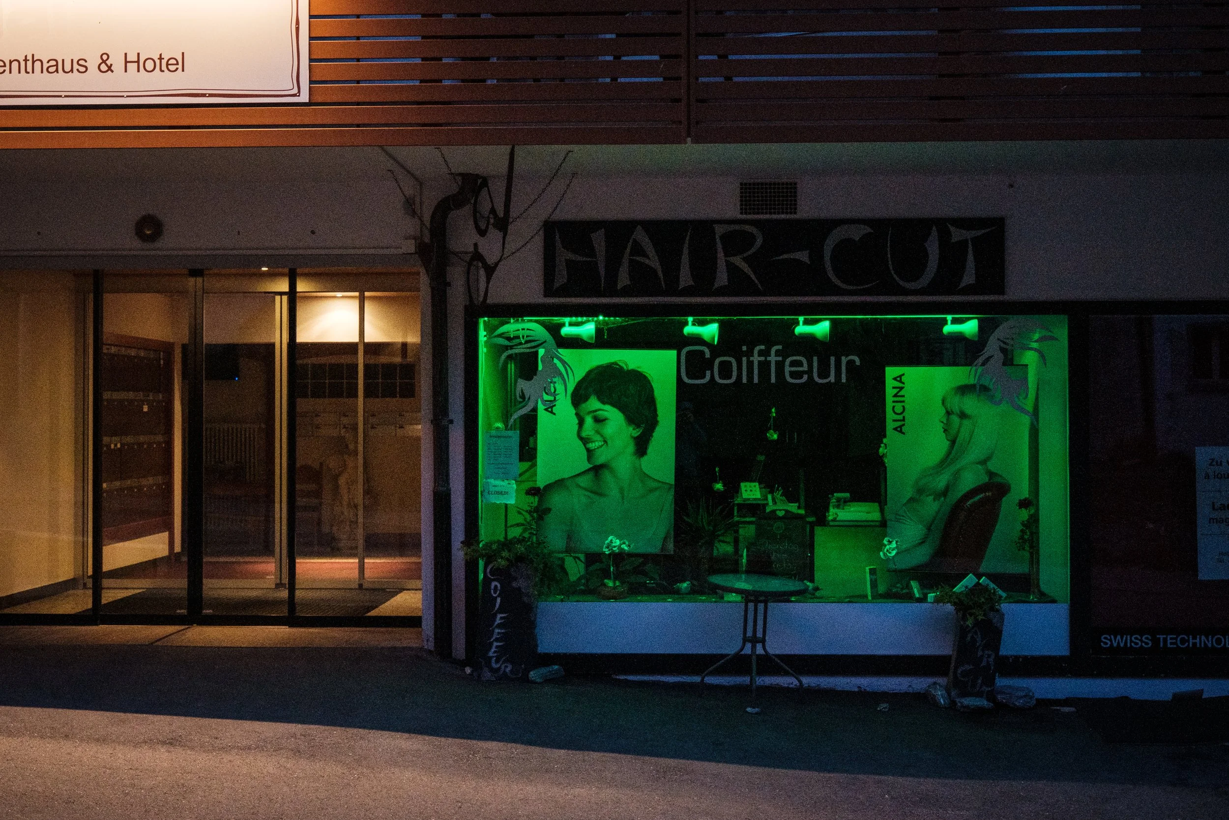 Schaufenster eines Friseursalons bei Nacht, mit grüner Beleuchtung und Fotos von Frauen mit unterschiedlichen Frisuren.