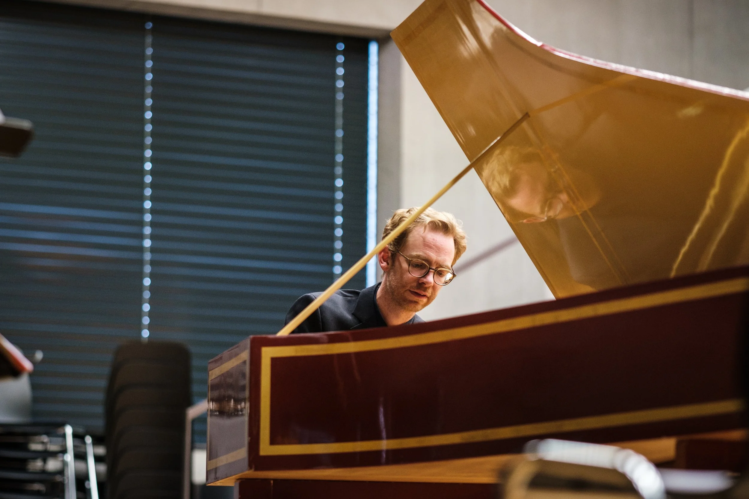 Ein Mann mit Brille und dunklem Anzug spielt auf einem alten, braunen Klavier in einem Raum mit blauen Fensterläden.