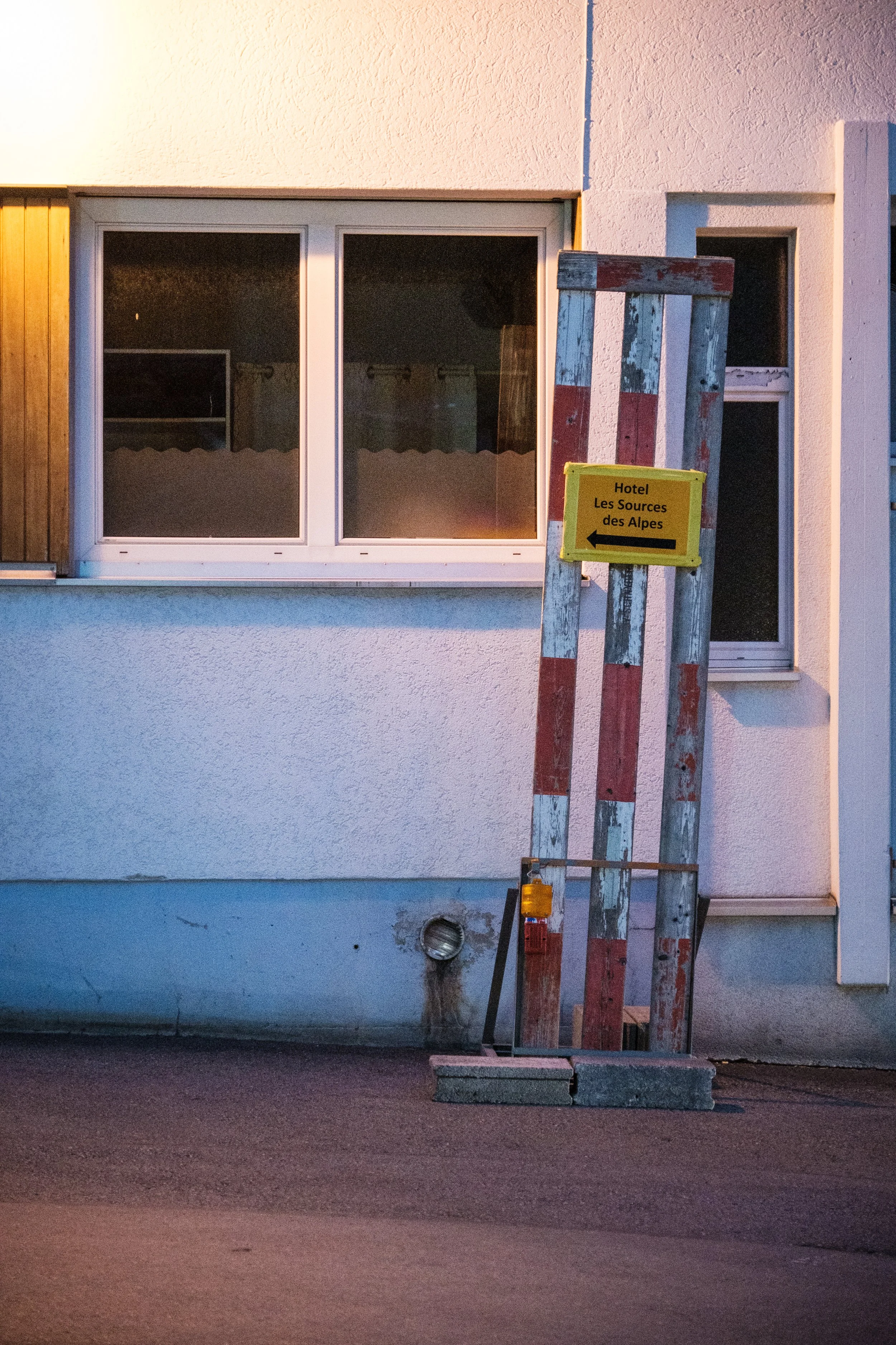 Zurückhaltende Szene eines weißen Gebäudes mit einem Fenster, einem alten Holzschild und einer Baustellenabsperrung mit gelbem Hinweisschild, das auf das Hotel Les Sources des Alpes zeigt, bei Dämmerung.