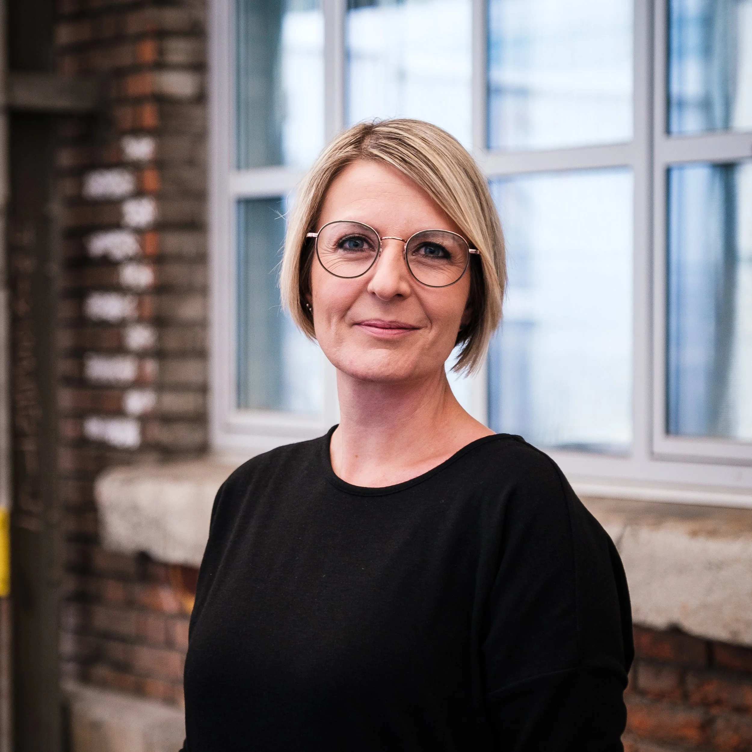 Porträt einer Frau mit kurzem blonden Haar, Brille und schwarzem Oberteil vor einer Ziegelmauer und Fenstern.