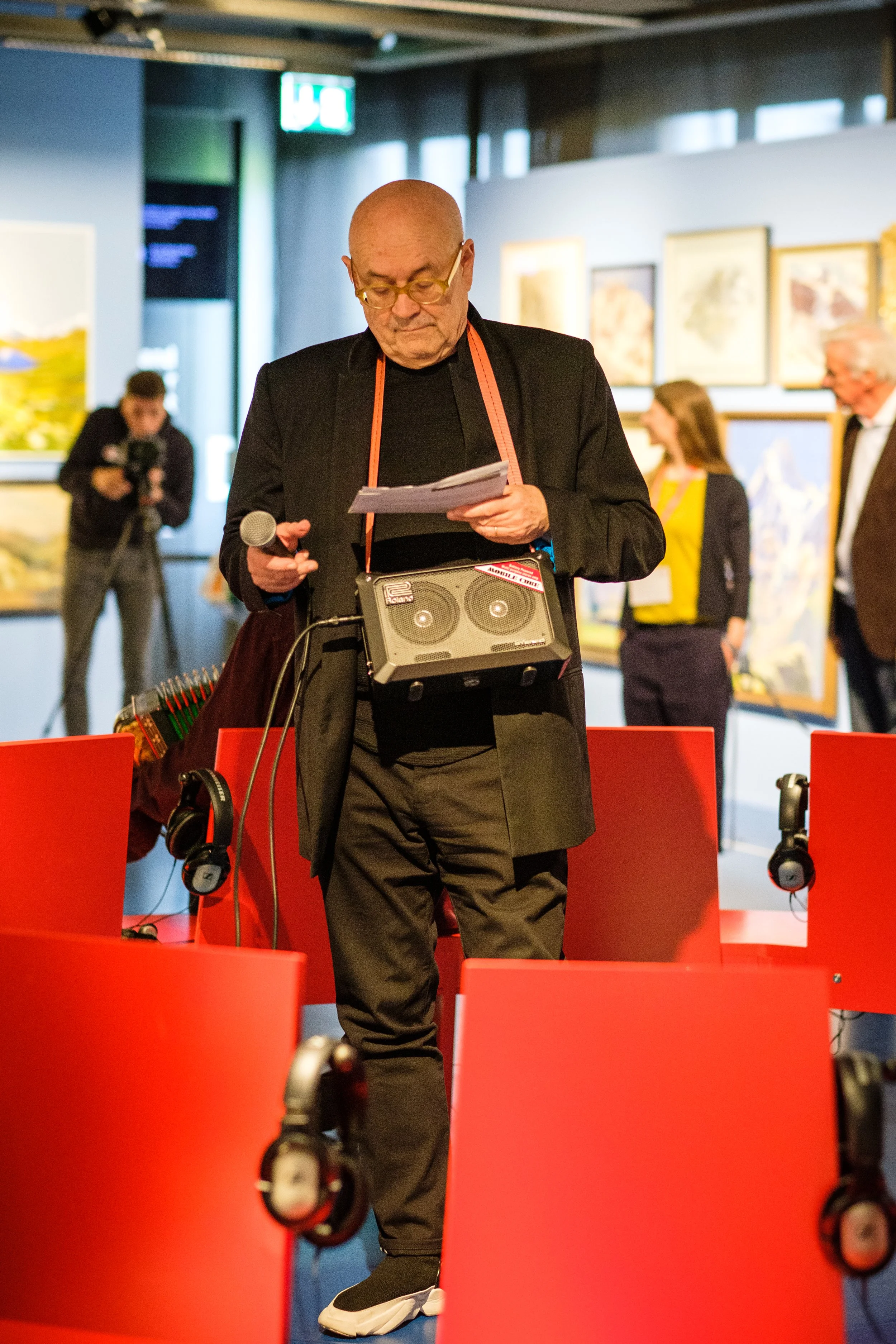Ein älterer Mann in schwarzer Jacke trägt ein Hörgerät und liest Notizen, während er in einem Kunstmuseum steht. Er hat eine kleine Lautsprecherbox um den Hals, und im Hintergrund sind weitere Personen sowie gerahmte Gemälde an der Wand zu sehen.