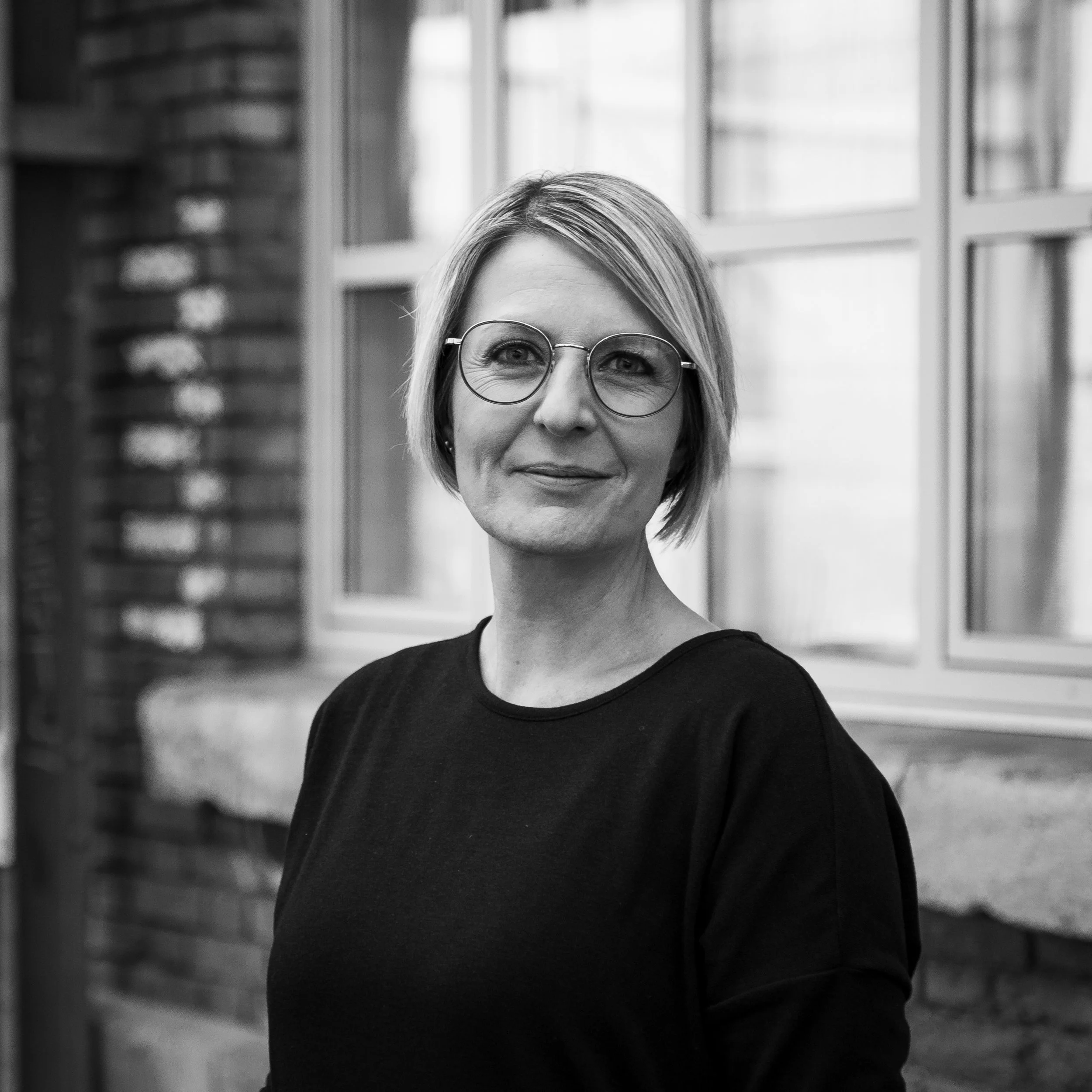 Schwarz-weiß-Foto einer Frau mit kurzen, glatten Haaren, Brille und einem schwarzen Pullover vor einem Fenster und einer Ziegelwand.