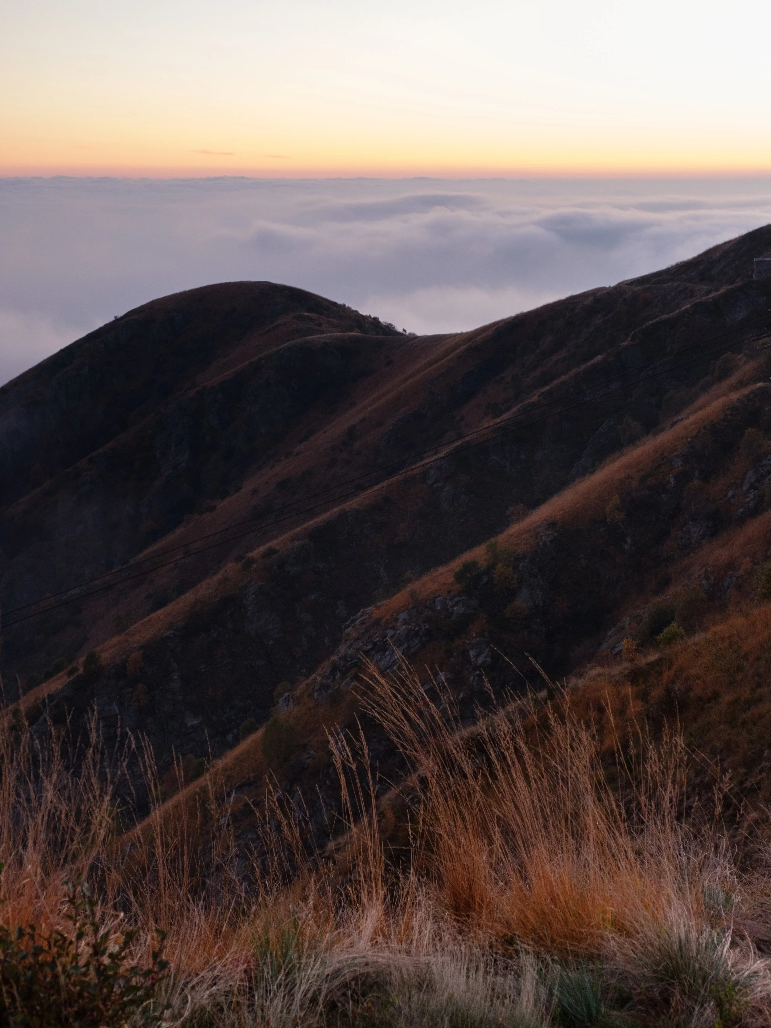 Berglandschaft bei Sonnenuntergang, mit Hügeln, hohen Gräsern im Vordergrund und Wolken unterhalb der Berge.