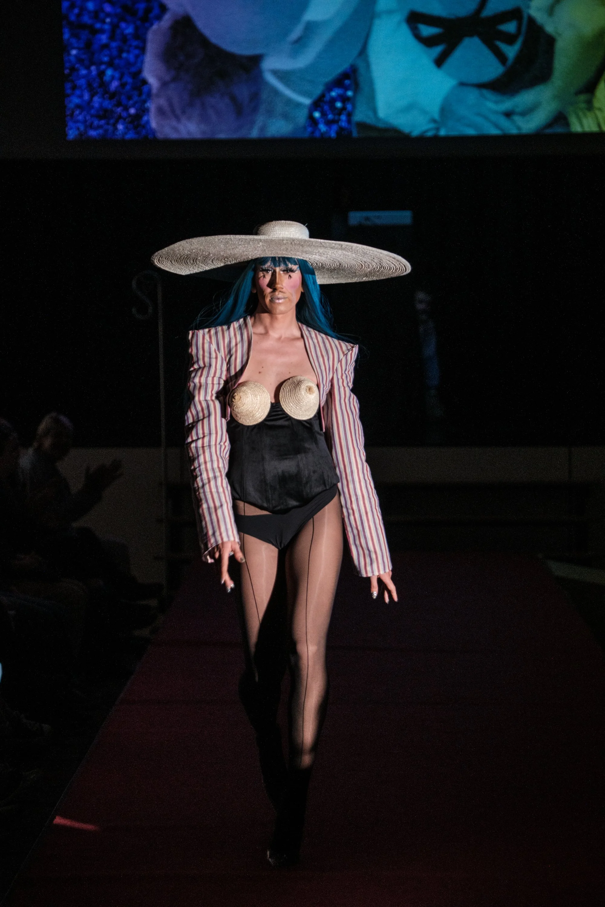 Model läuft auf Laufsteg, trägt großen Strohhut, gestreiftes Jackett, schwarzes Top mit angenähten Muscheln, schwarzen Shorts und durchsichtige Strümpfe.