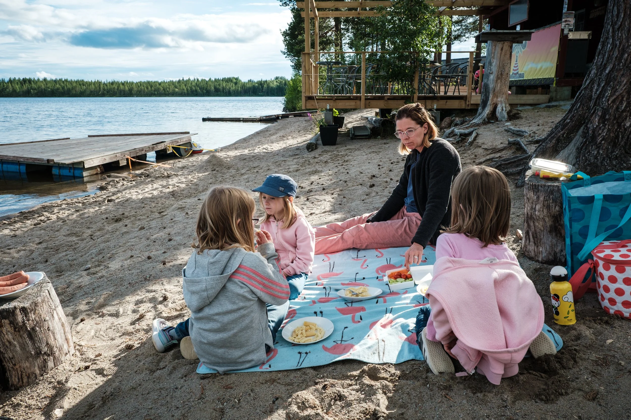 Familie, die am Strand ein Picknick auf einer Flamingo-dekorierten Decke unter Bäumen macht, mit Wasser, Steganlage und Boot im Hintergrund.