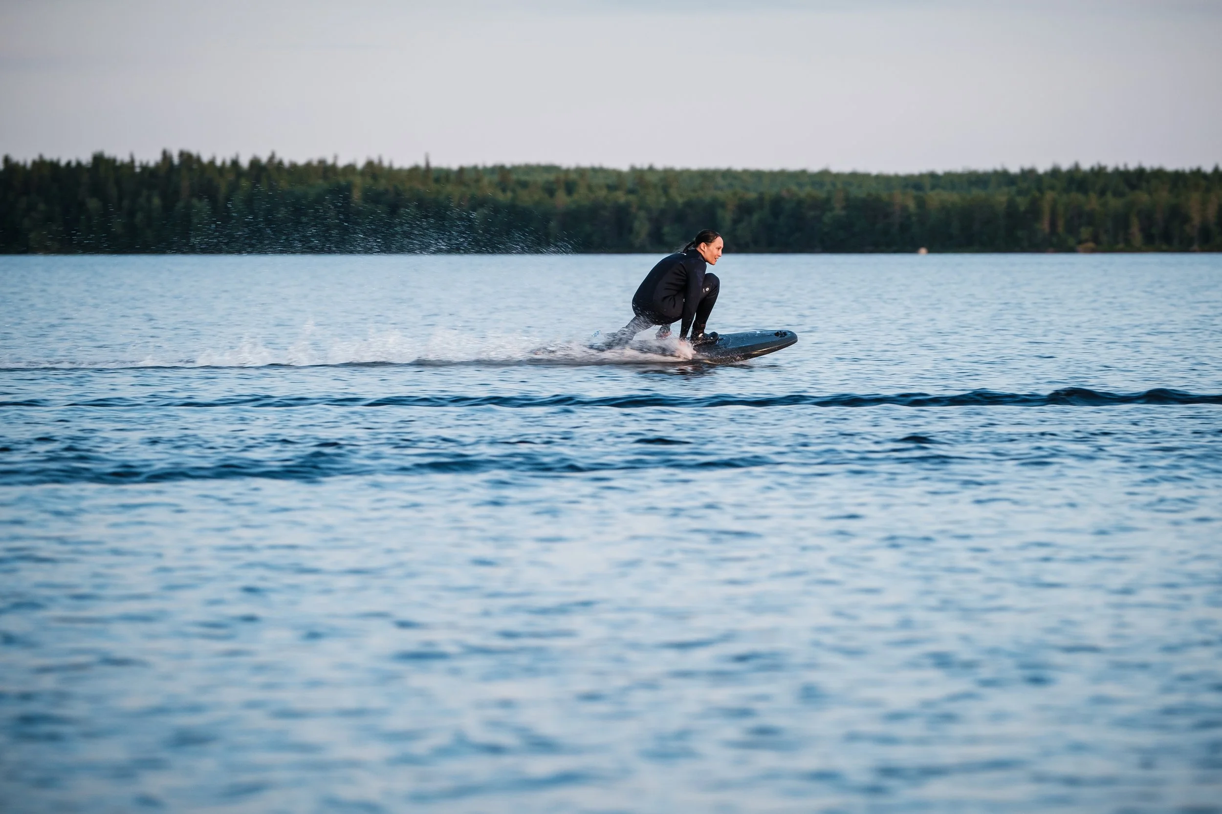 Frau, die auf einem Wasserboard auf einem See surft, mit Wald im Hintergrund.