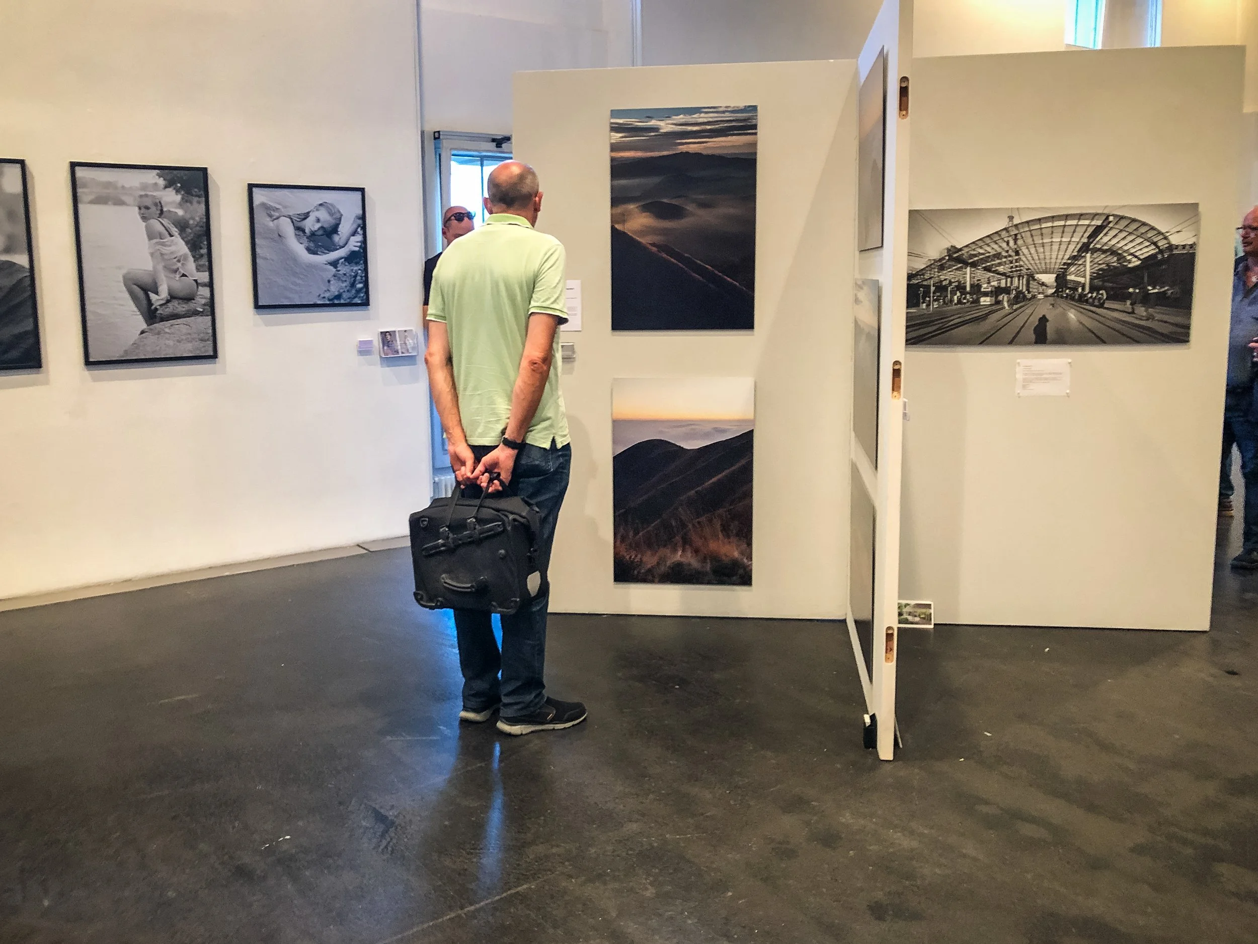 Ein Mann mit schwarzer Tasche betrachtet Kunstfotografien in einer Galerie, andere Besucher sind im Hintergrund zu sehen.
