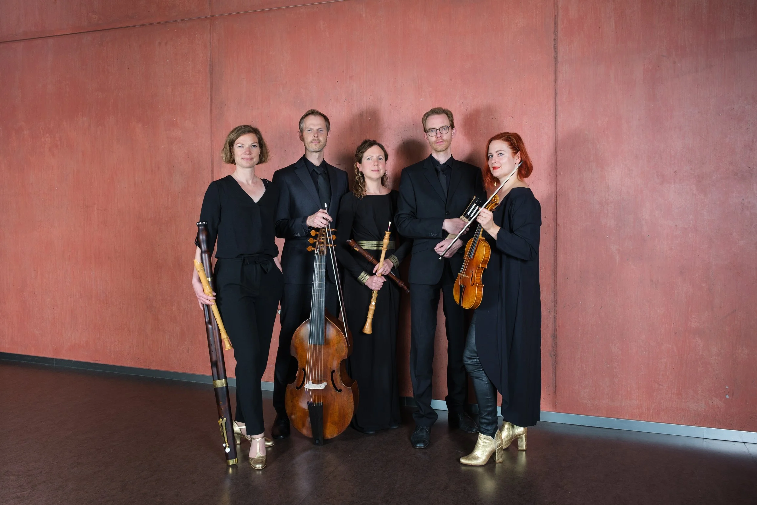 Fünf Musiker stehen vor einer roten Wand, halten verschiedene Instrumente, tragen schwarze Kleidung, einige mit goldfarbenen Details, und schauen in die Kamera.