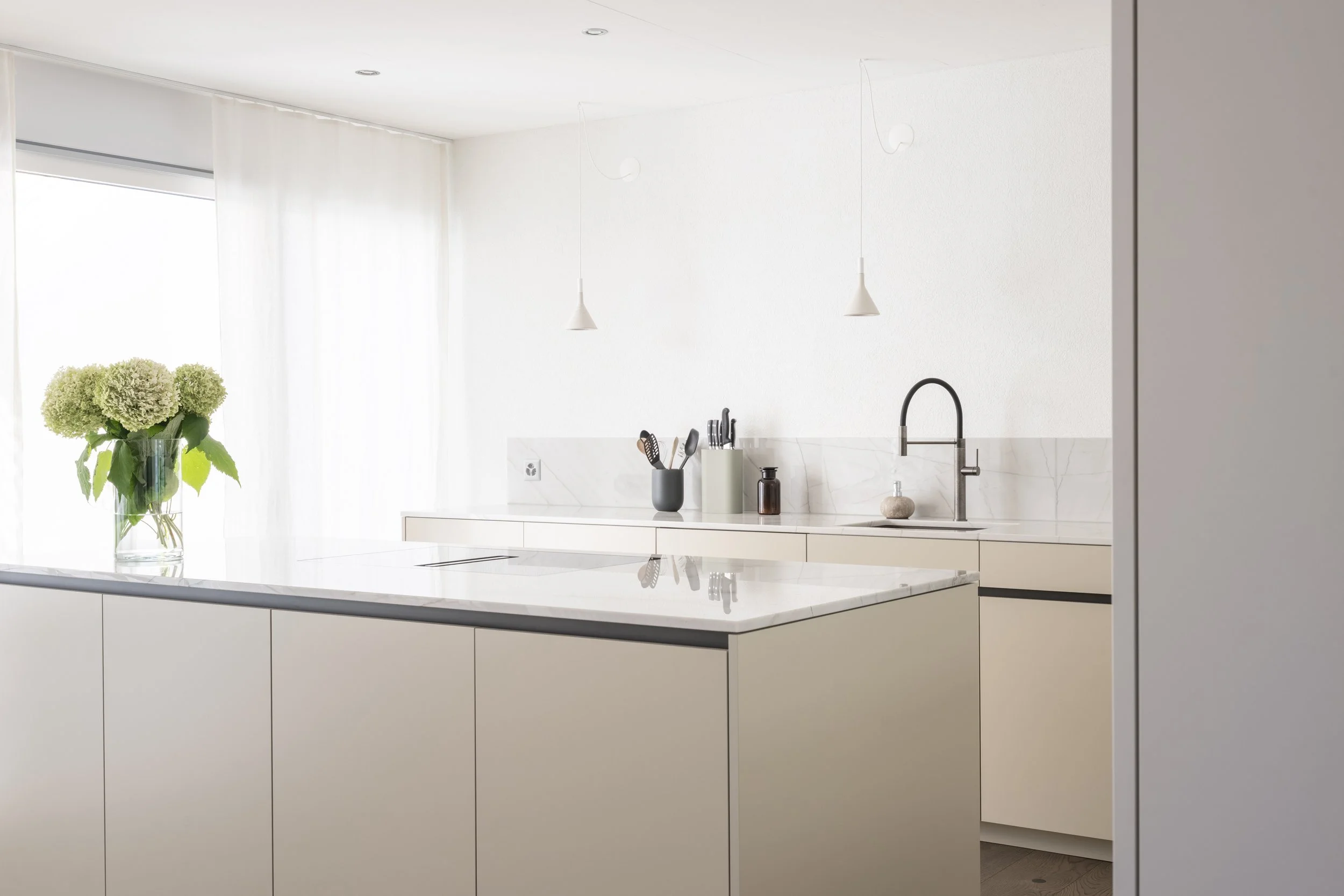 Moderne weiße Küche mit minimalistischem Design, Kochinsel mit gekalkter Oberfläche, Blumenstrauß in Glasvase auf der linken Seite, schlichte Armaturlänge, Hänge-Lampen, Wasserhahn an der Spüle, Küchenutensilien in Behältern, helle Gardinen vor Fenst