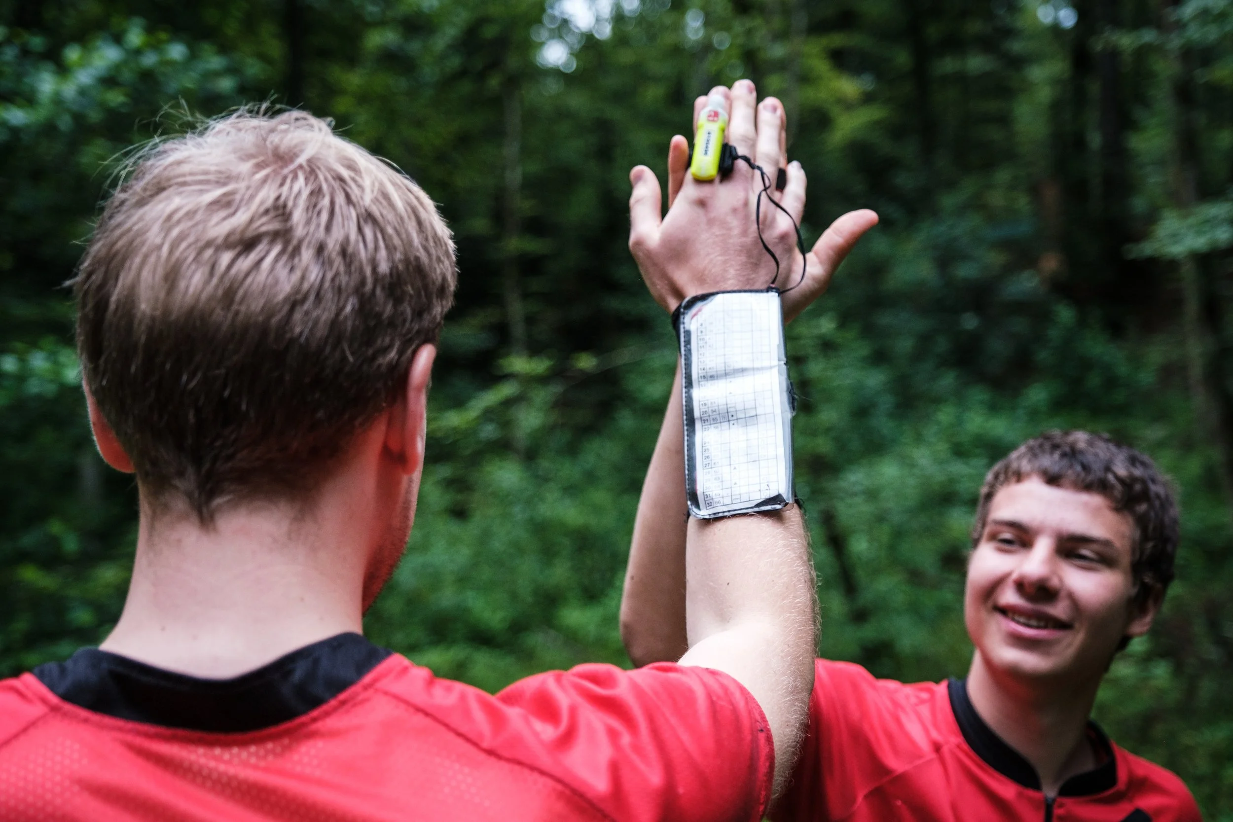 Zwei junge Männer in roten Trikots im Wald, einer zeigt auf seinen Arm mit einem Fitness-Tracker, während der andere lächelt.