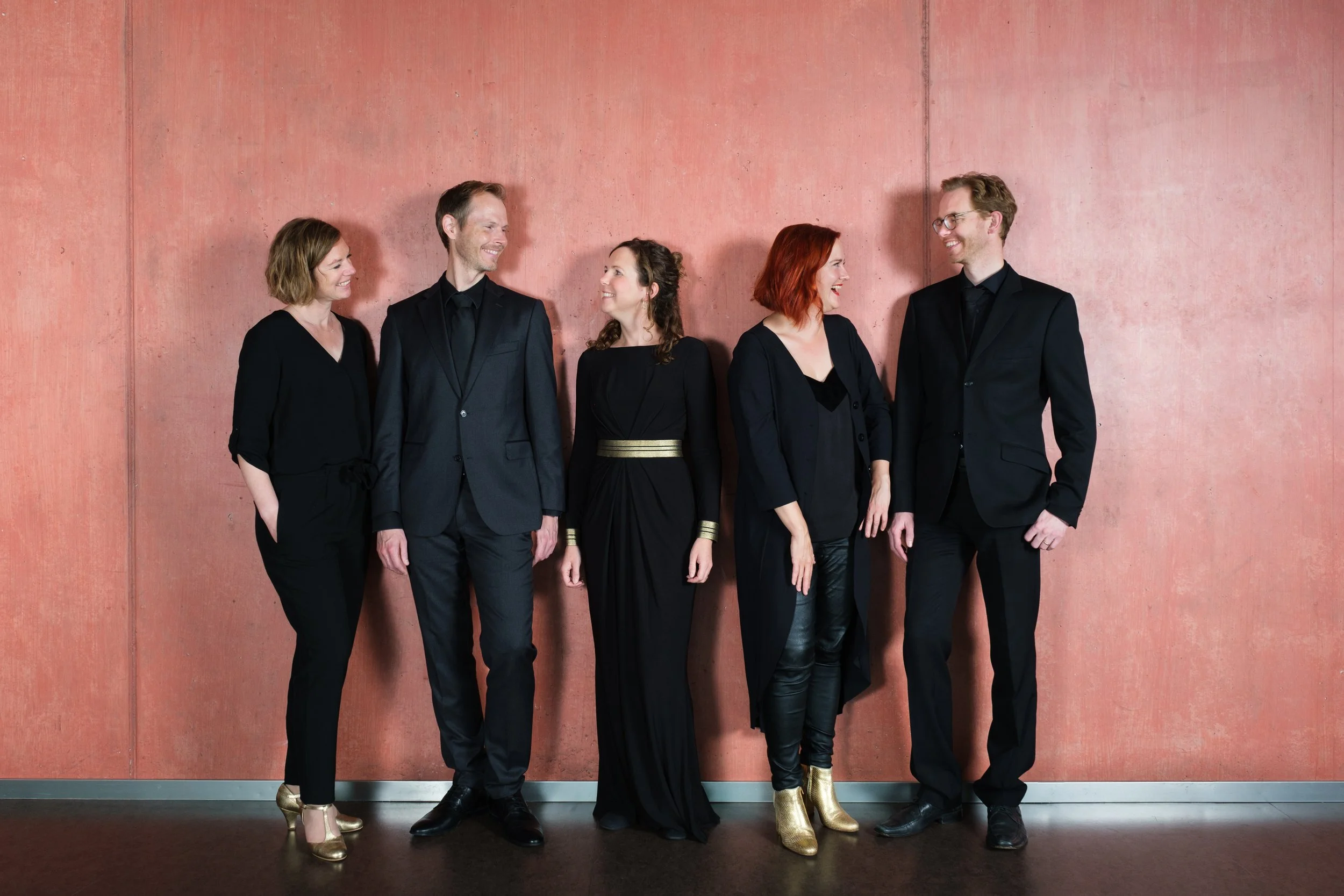 Fünf Menschen in schwarzen Outfits, die nebeneinander vor einer roten Wand stehen und lachen.