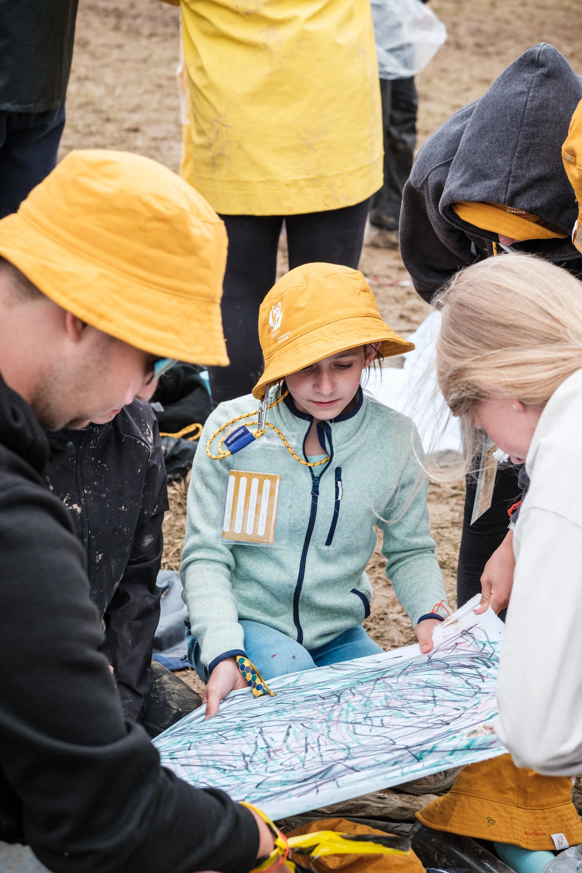 Kinder und Erwachsene sitzen im Sand, betrachten gemeinsam ein großes, buntes, von Kinderhand gestaltetes Kunstwerk.