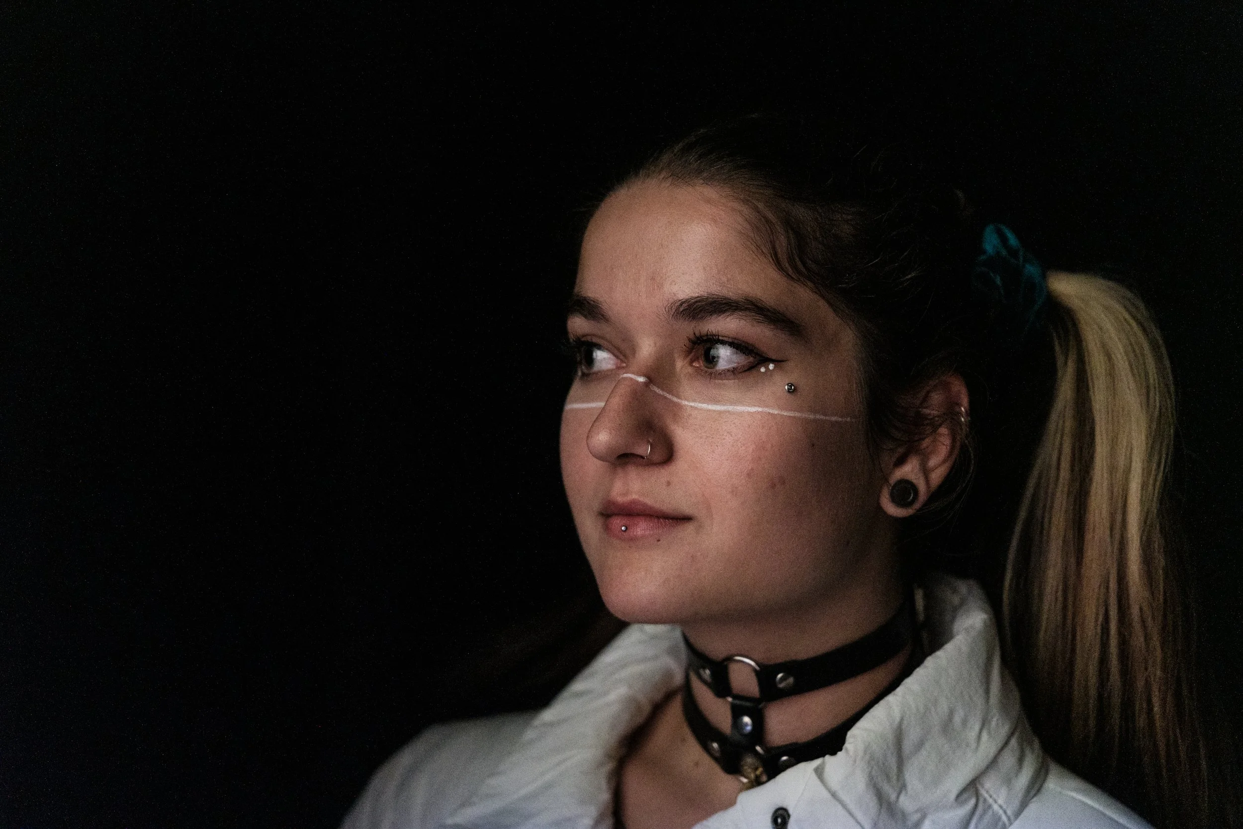 Junge Frau mit Piercings und Choker-Kette blickt seitlich vor einem schwarzen Hintergrund.