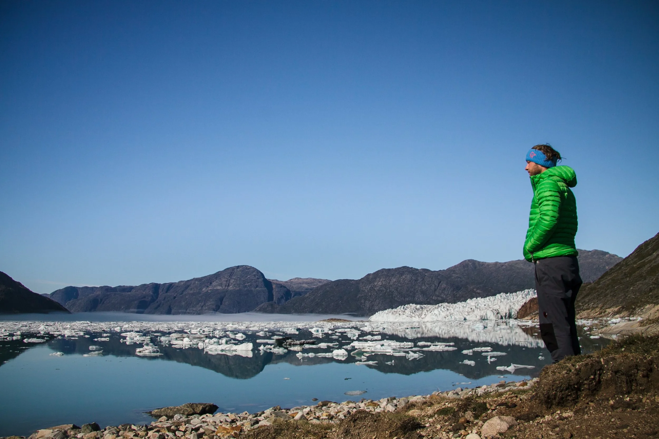 Mann in grünem Jacke und blauer Stirnband steht am Rand eines ruhigen Sees mit Eis und Bergen im Hintergrund, bei sonnigem Himmel.
