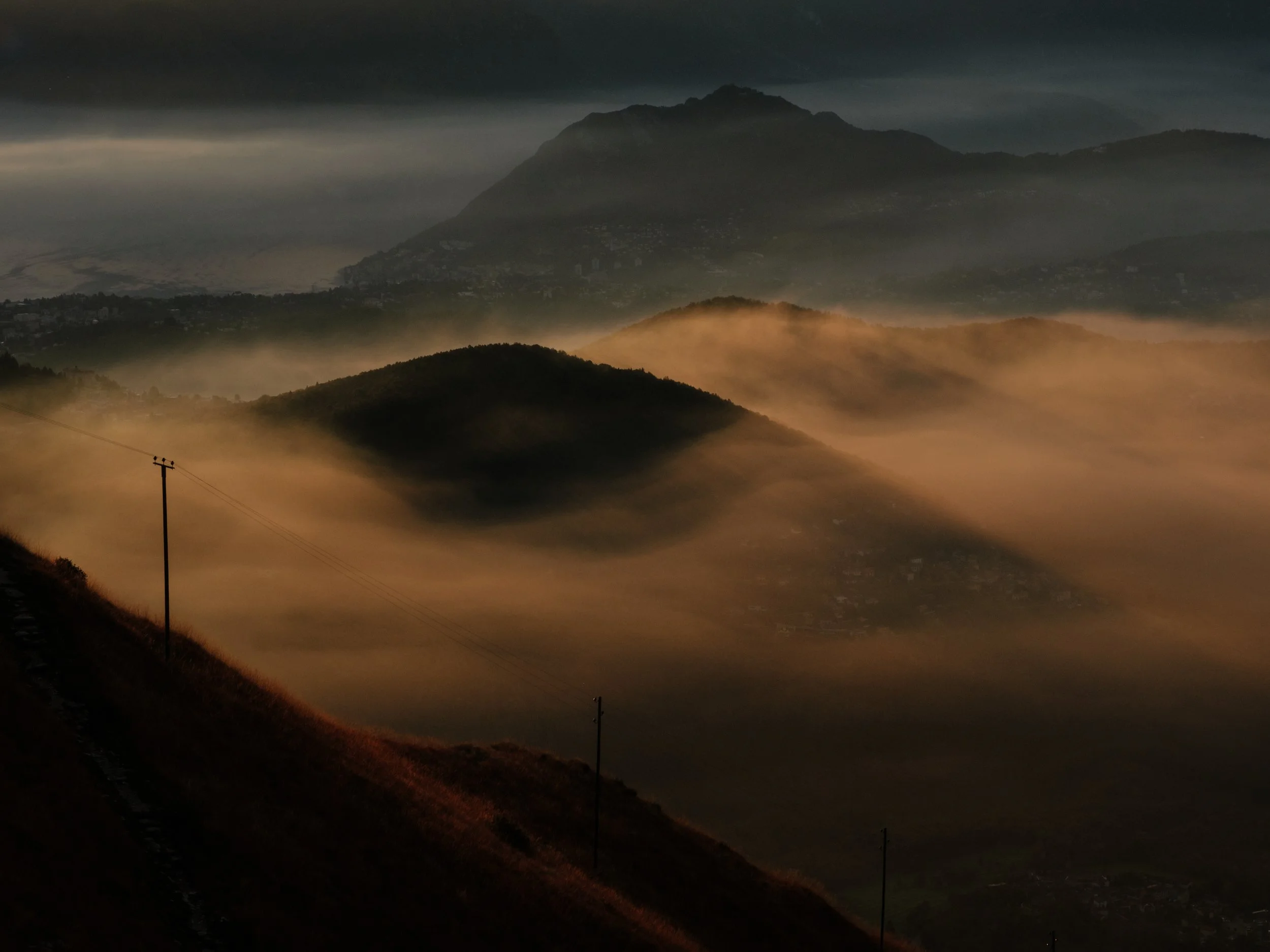 Berglandschaft im Nebel bei Sonnenauf- oder untergang mit Hügeln, Strommasten und Bergen im Hintergrund.