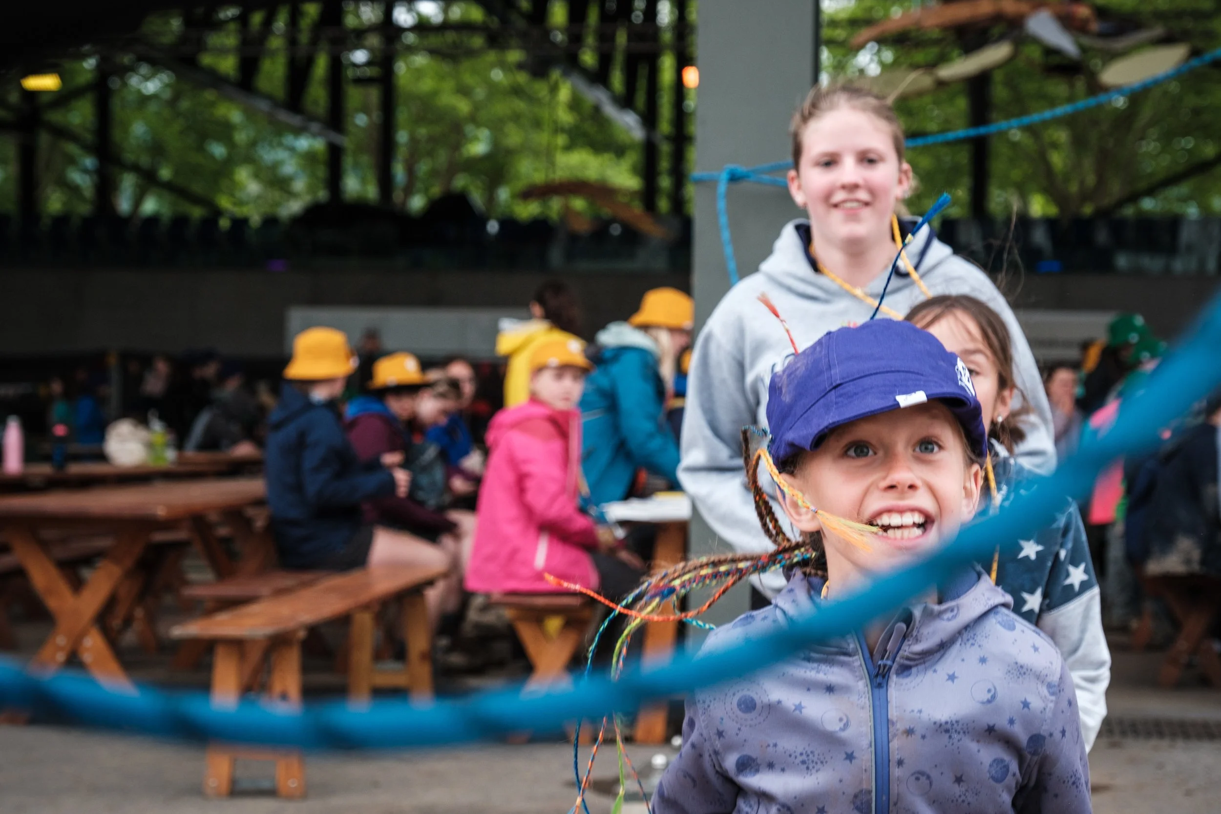 Kinder beim Kinder- und Jugendzeltlager, einige sitzen an Tischen und andere stehen, im Hintergrund sind Bäume und eine Gebäudehalle sichtbar.