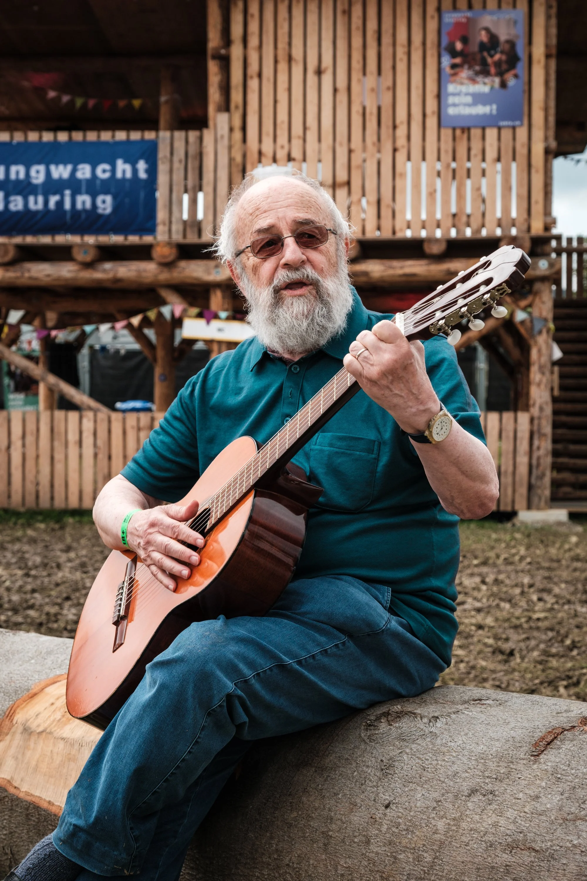 Ein älterer Mann mit weißem Bart sitzt auf einem Baumstamm und spielt Gitarre. Im Hintergrund ist eine Holzstruktur mit Schildern und Dekorationen.