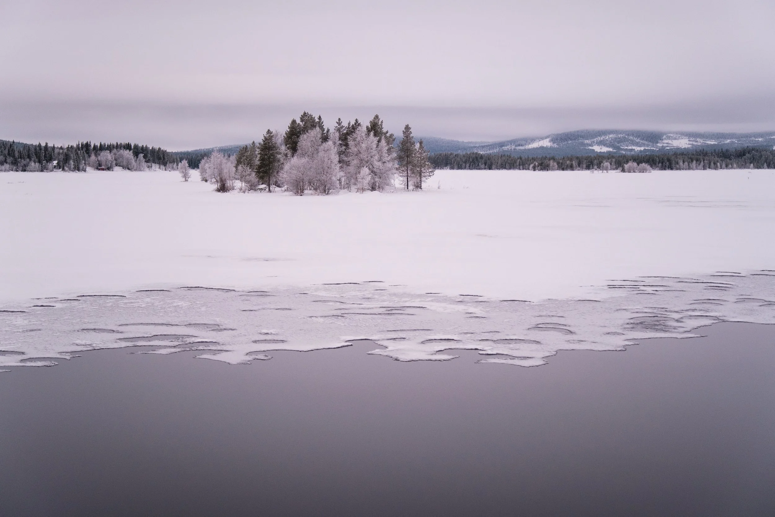Eis- und Schneelandschaft mit Baumgruppe auf einer Insel im gefrorenen See, bewölkter Himmel, schneebedeckte Wälder im Hintergrund.