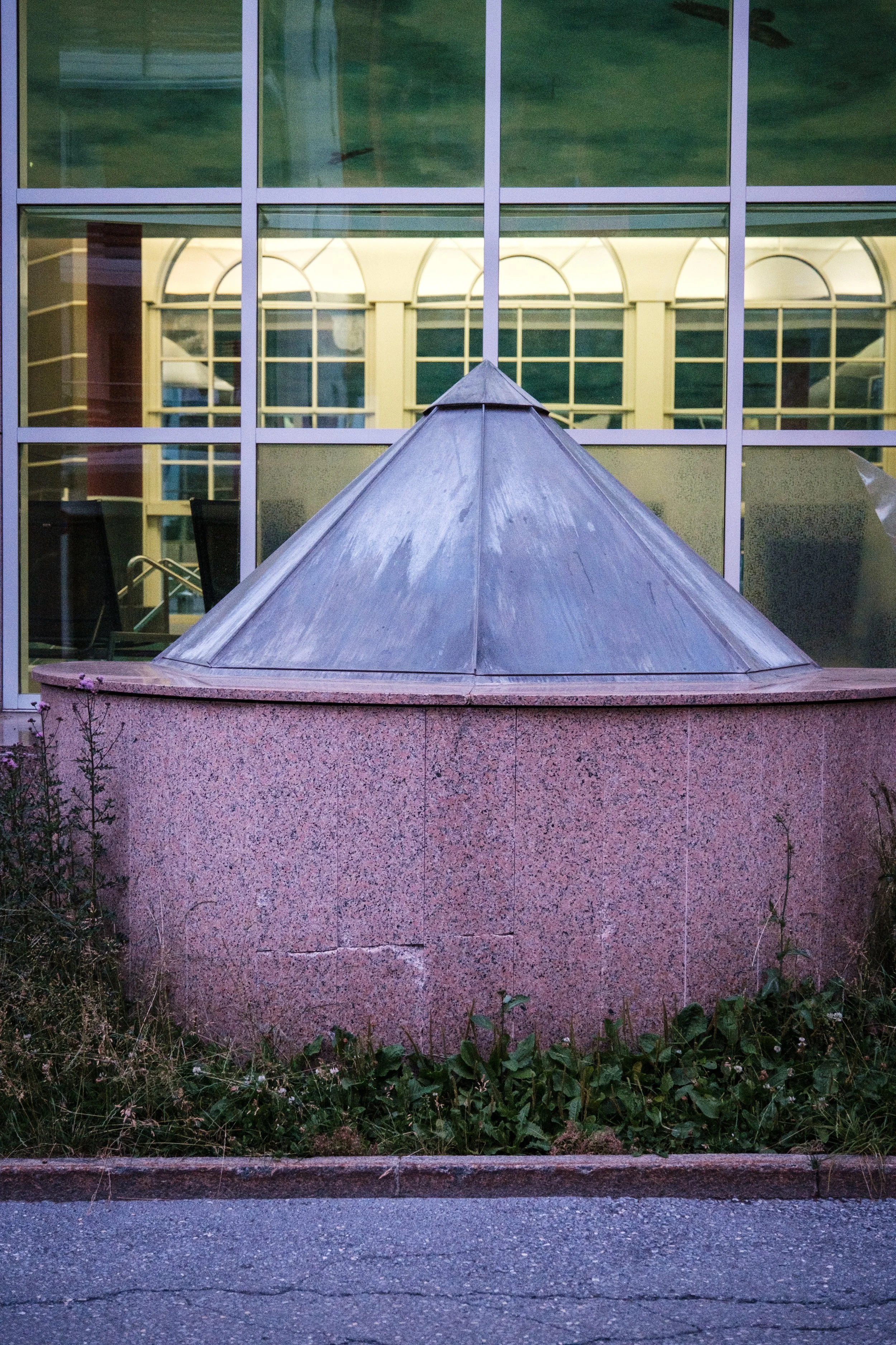 Ein rundes, pinkfarbenes Brunnen mit kegelförmiger metallischer Abdeckung vor einem modernen Gebäude mit großen Fenstern.