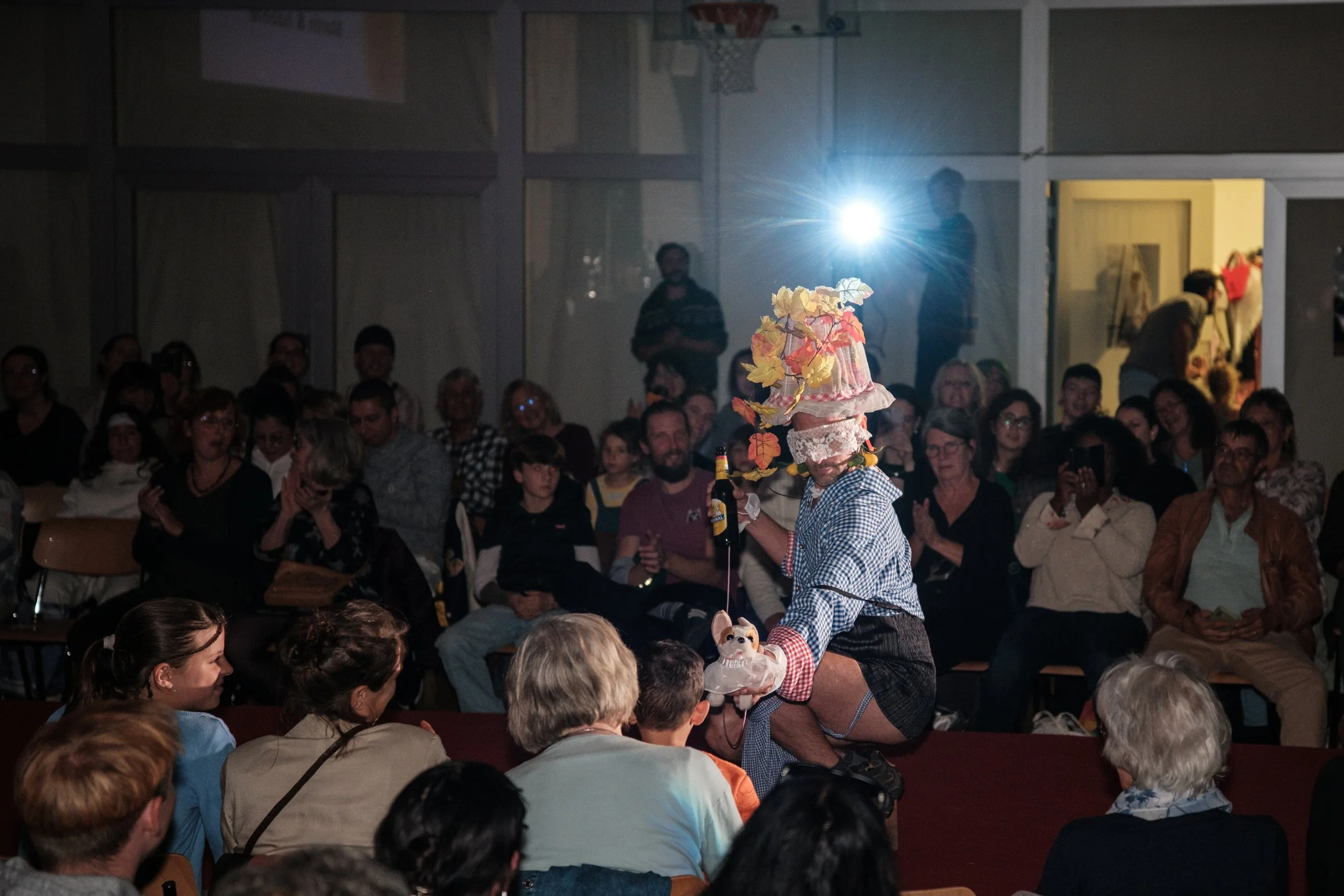 Ein Performer mit einem auffälligen Hut, der mit Blumen und Blättern geschmückt ist, bei einer Bühnenshow vor Publikum.