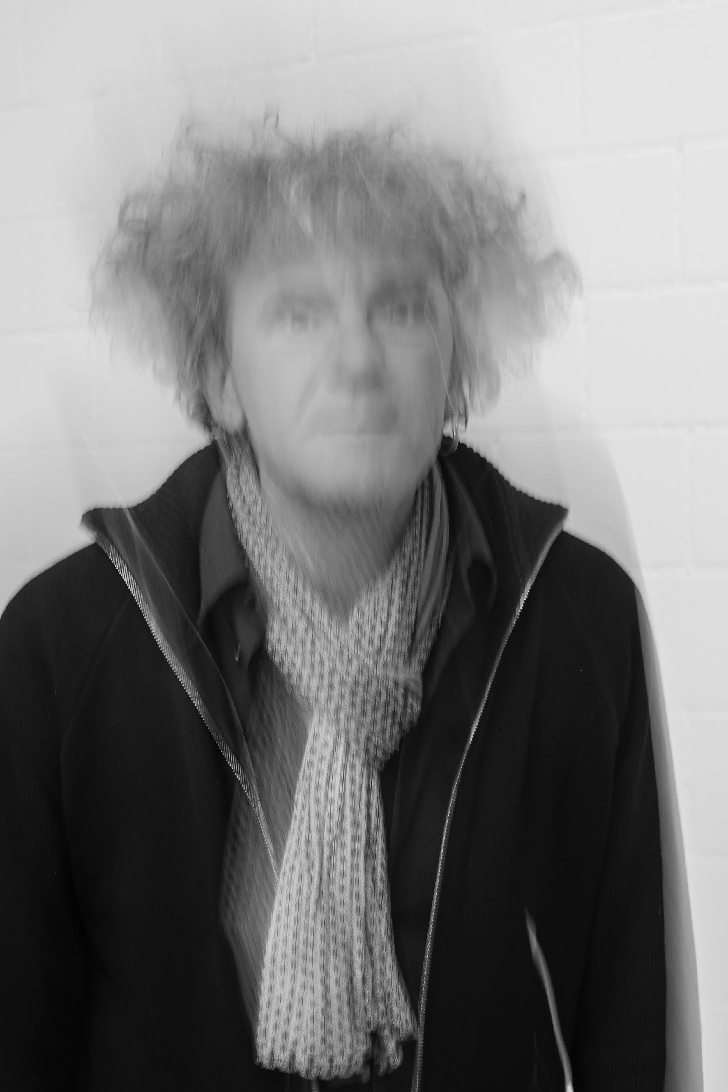 Schwarz-weiß-Fotografie einer Person mit lockigem, unordentlichem Haar vor einer weißen Wand, die eine dunkle Jacke, ein Hemd und einen Schal trägt.