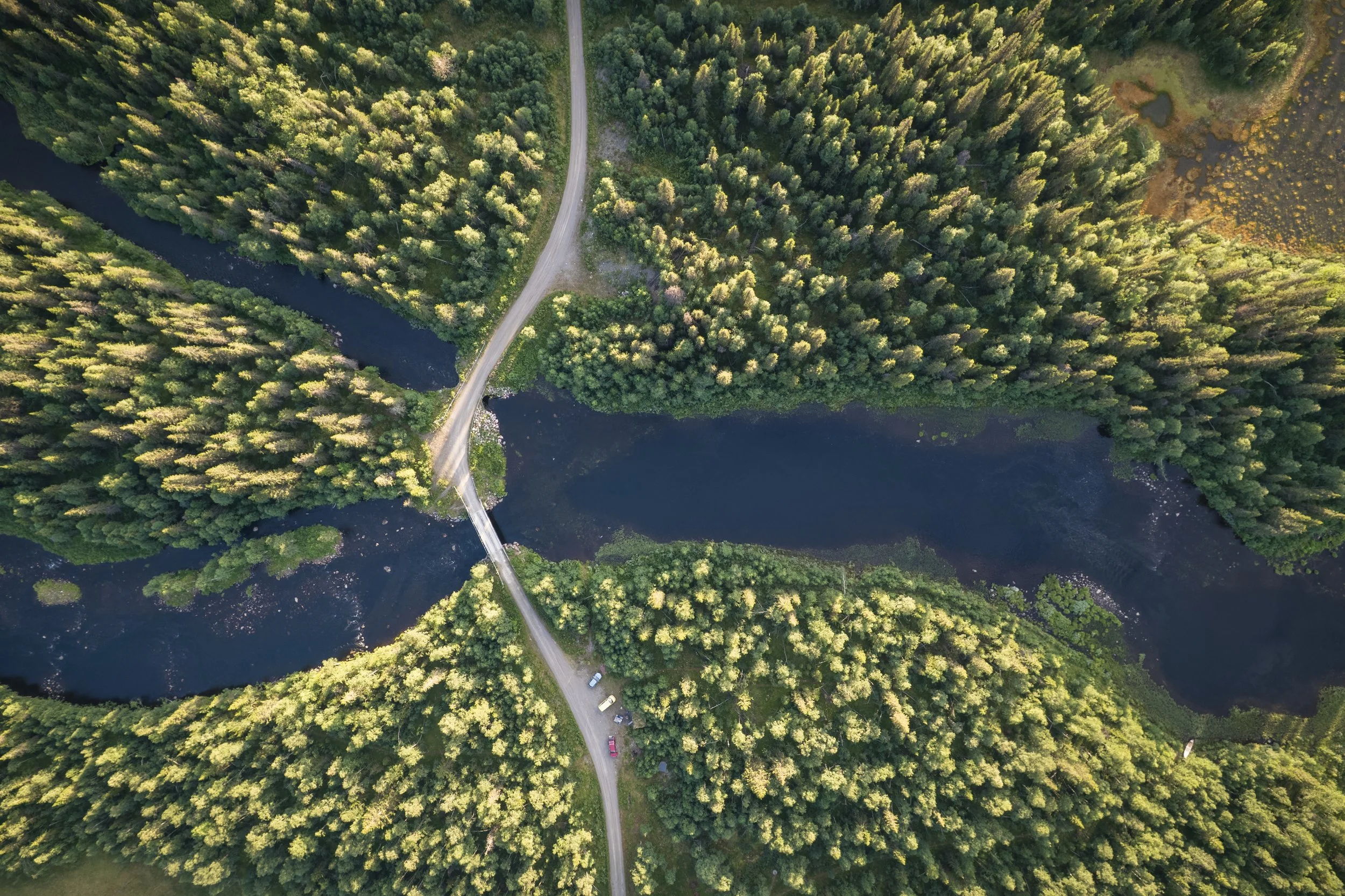 Luftaufnahme eines Flusses, der durch einen dichten, grünen Wald fließt, mit einer Brücke, die die beiden Seiten verbindet, und einem kleinen Parkplatz darunter.