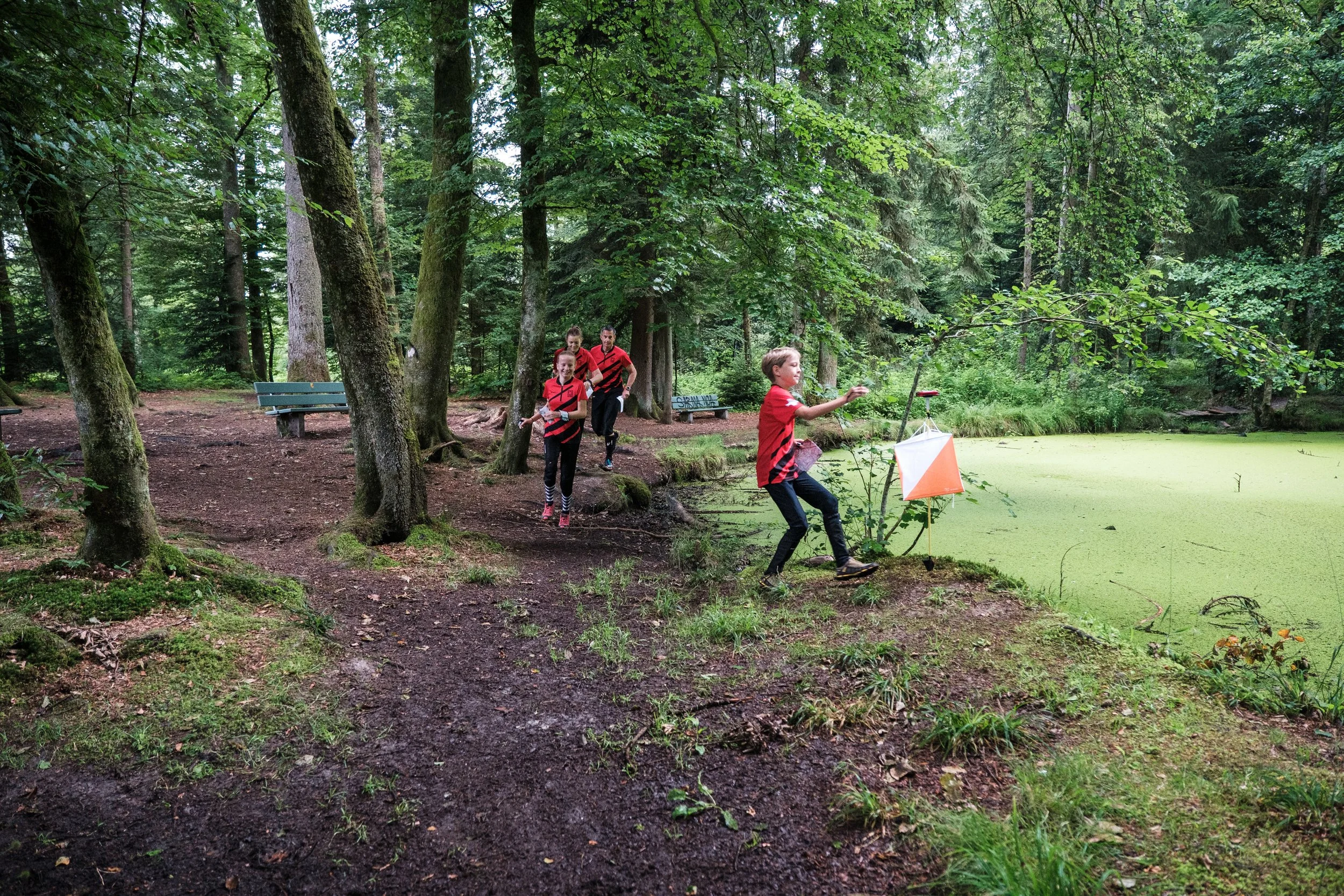 Vier Kinder in rot-schwarzen Trikots beim Spielen im Wald, eines wirft einen Gegenstand in den See, während die anderen zuschauen, umgeben von Bäumen und einem grünen See.
