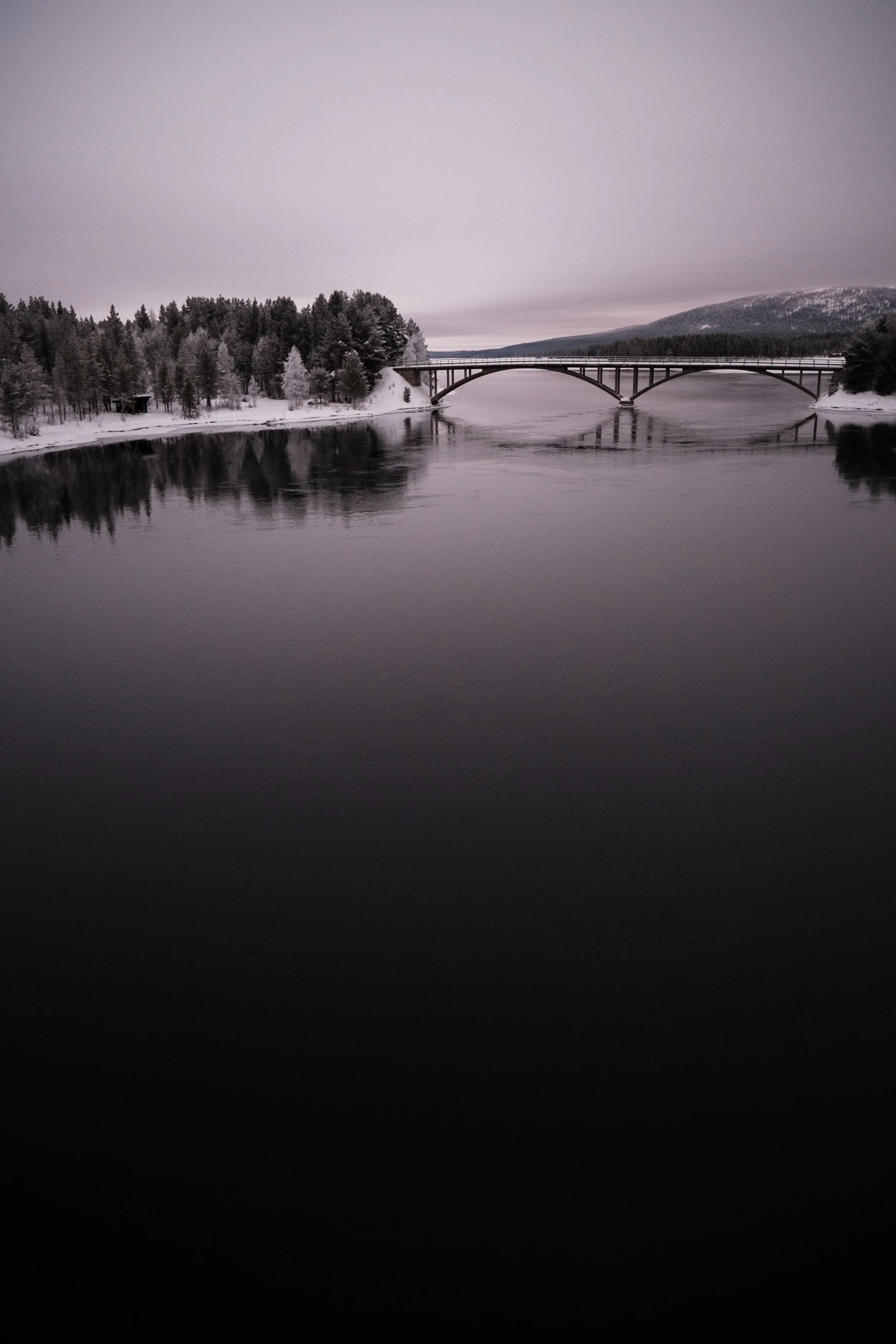 Schneebedecktes Land mit Brücke über einen ruhigen Fluss im Winter, bewölkter Himmel.